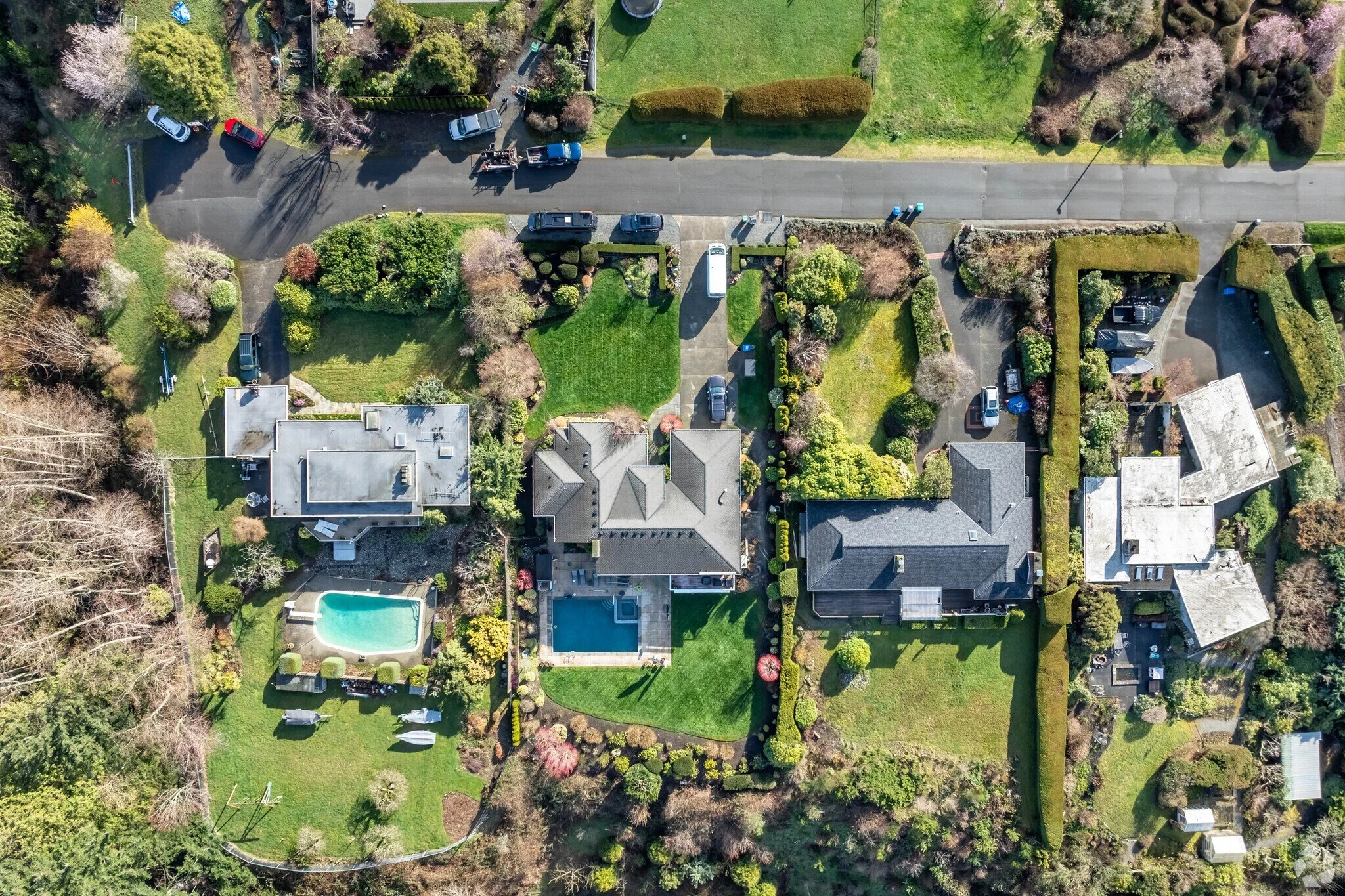 Aerial view of a residential neighborhood showing several houses with yards and swimming pools, trees, and parked cars along a street.