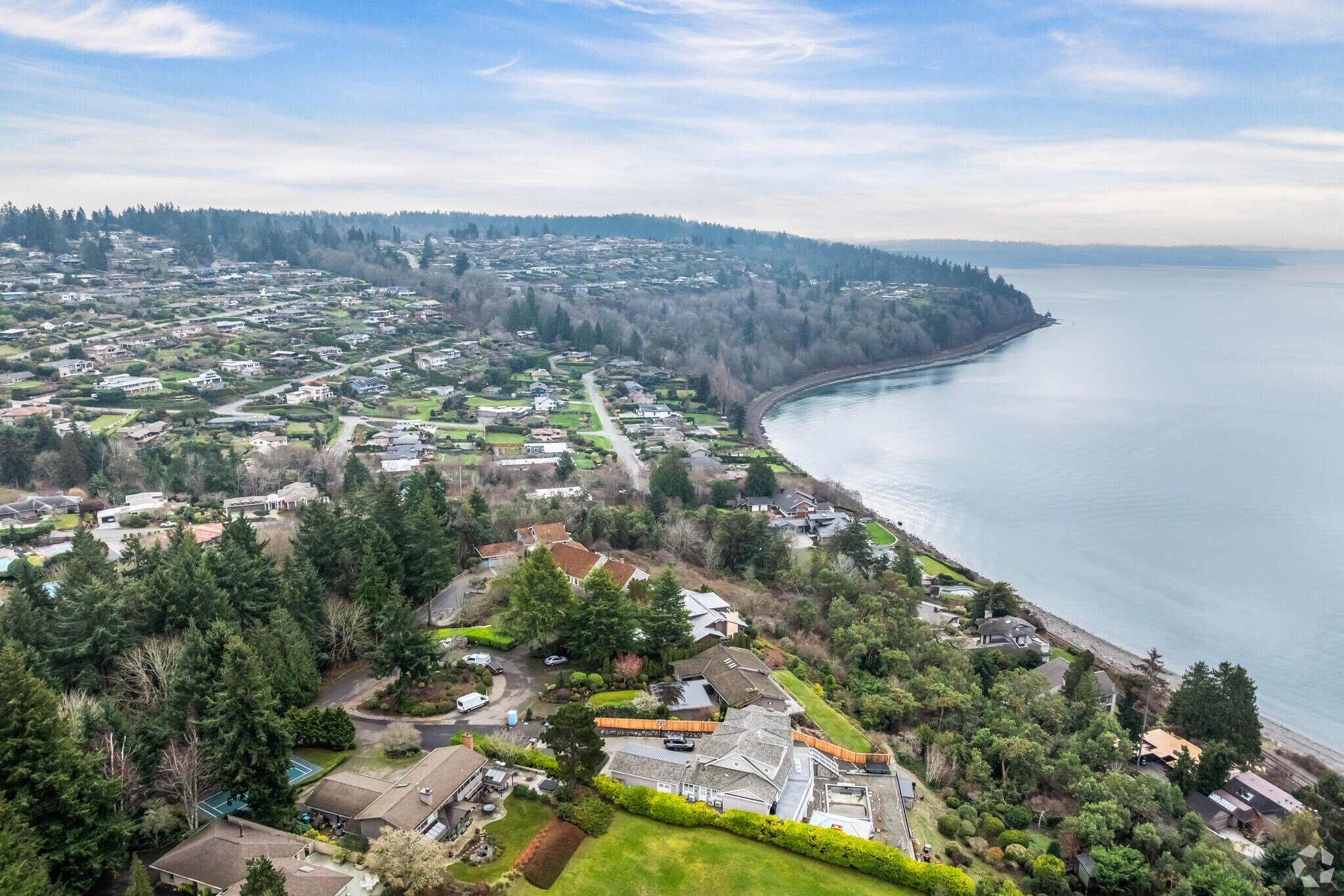 Aerial view of a coastal residential area with houses, trees, and a shoreline along a large body of water, under a cloudy sky.