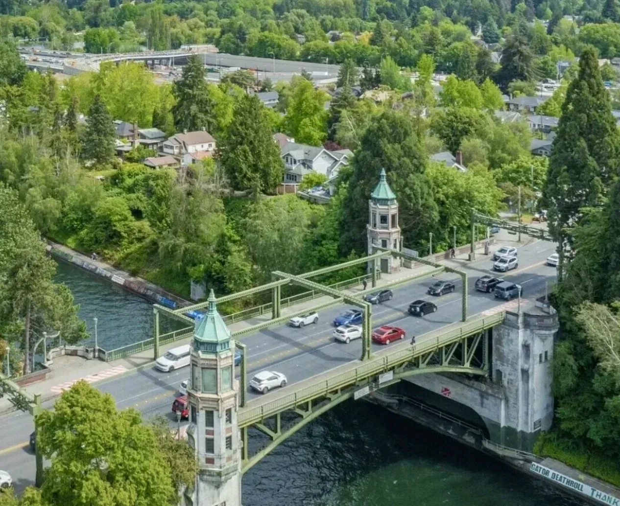 An aerial view of a bridge crossing a river, surrounded by lush green trees and a residential neighborhood with houses and roads in the background.