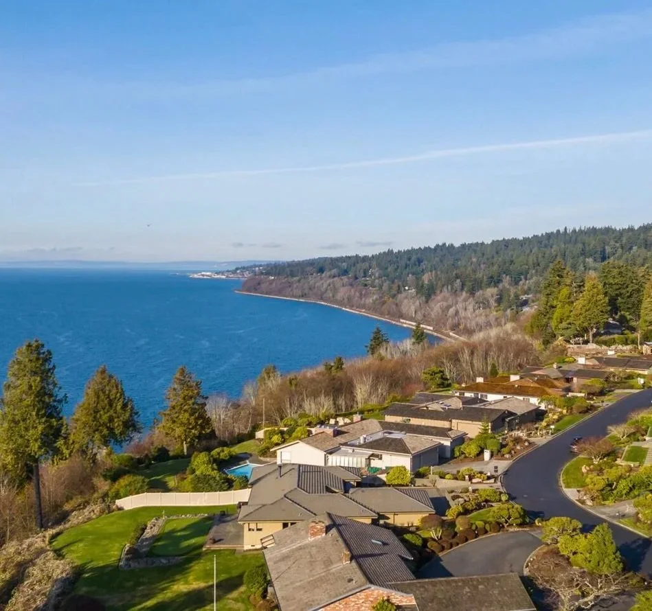 Aerial view of a coastal residential neighborhood with houses, trees, winding roads, and a large body of water in the background under a clear blue sky.