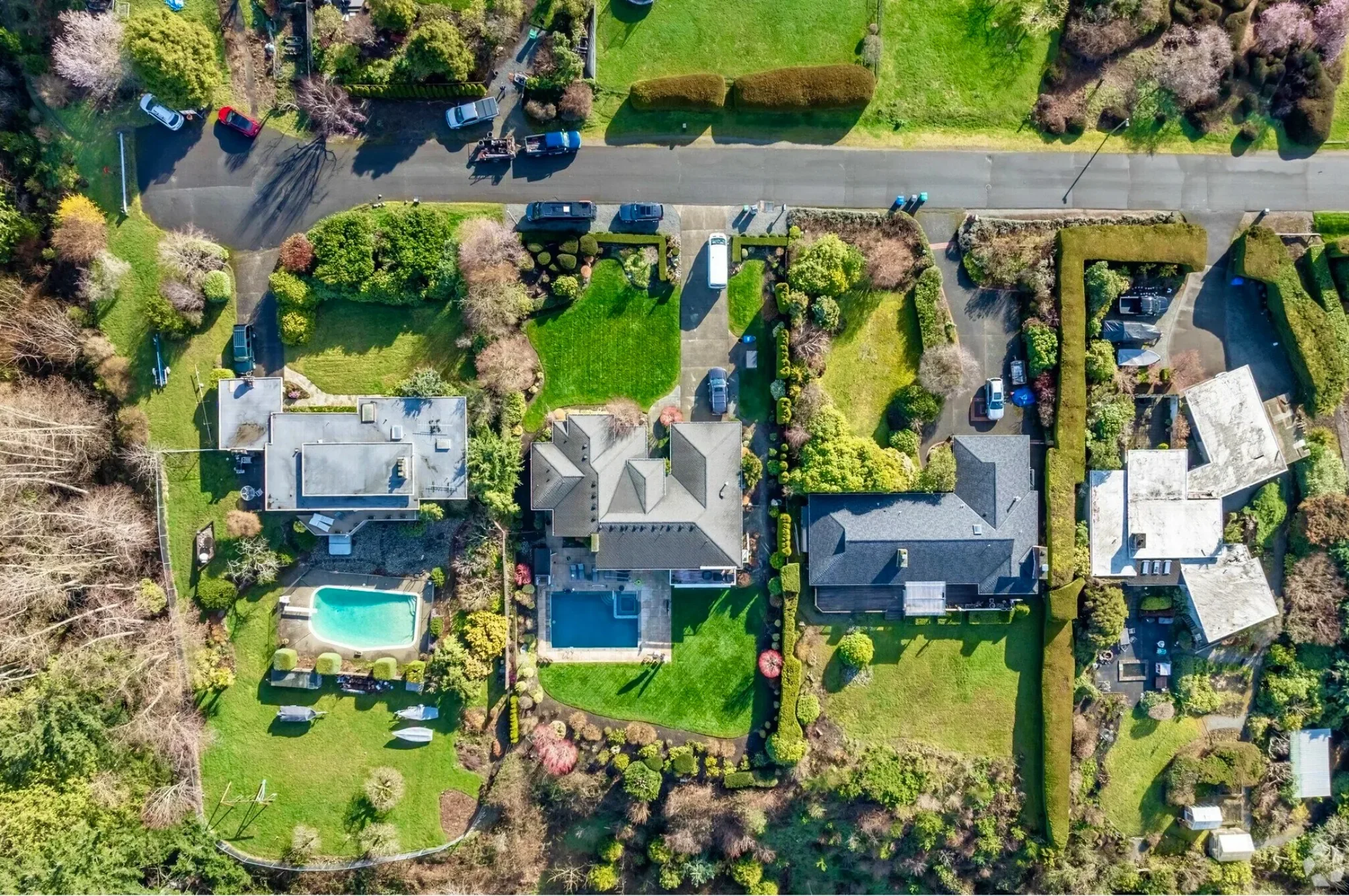 Aerial view of a residential neighborhood showing several houses with yards and swimming pools, trees, and parked cars along a street.