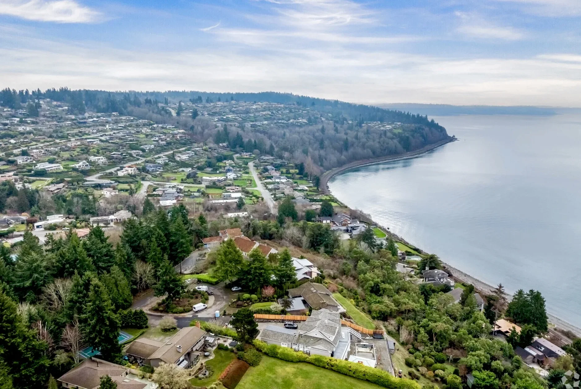 Aerial view of a coastal residential area with houses, trees, and a shoreline along a large body of water, under a cloudy sky.