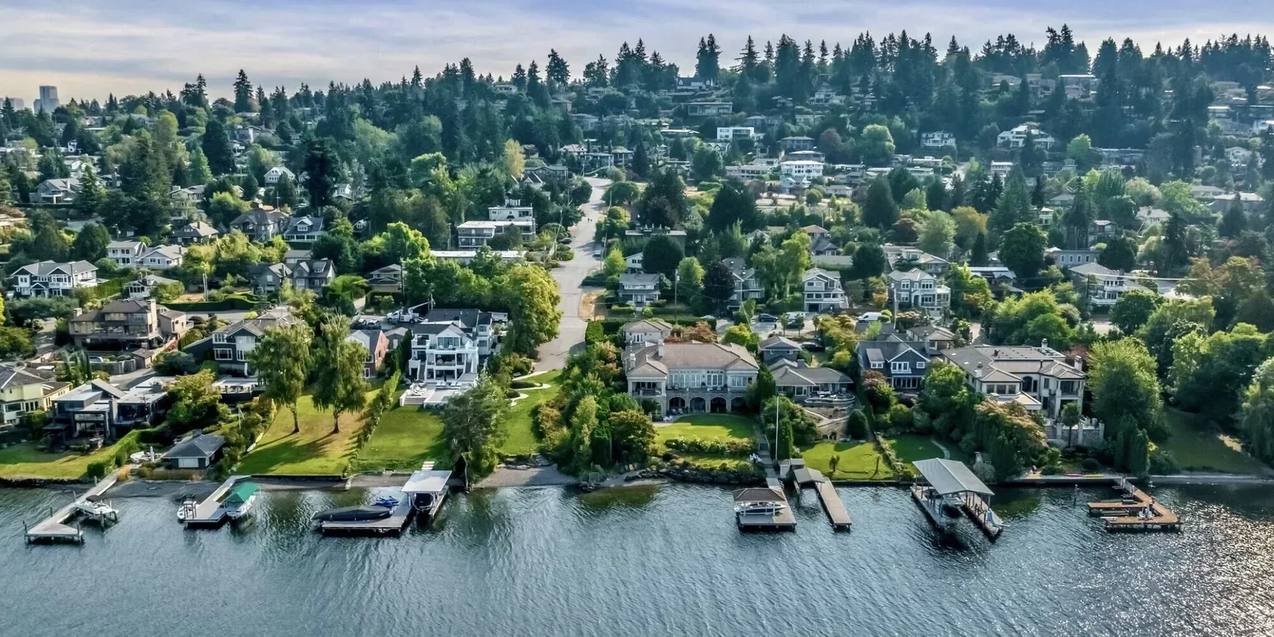 Aerial view of waterfront residential neighborhood with houses and docks along a river, surrounded by green trees and hilly terrain.