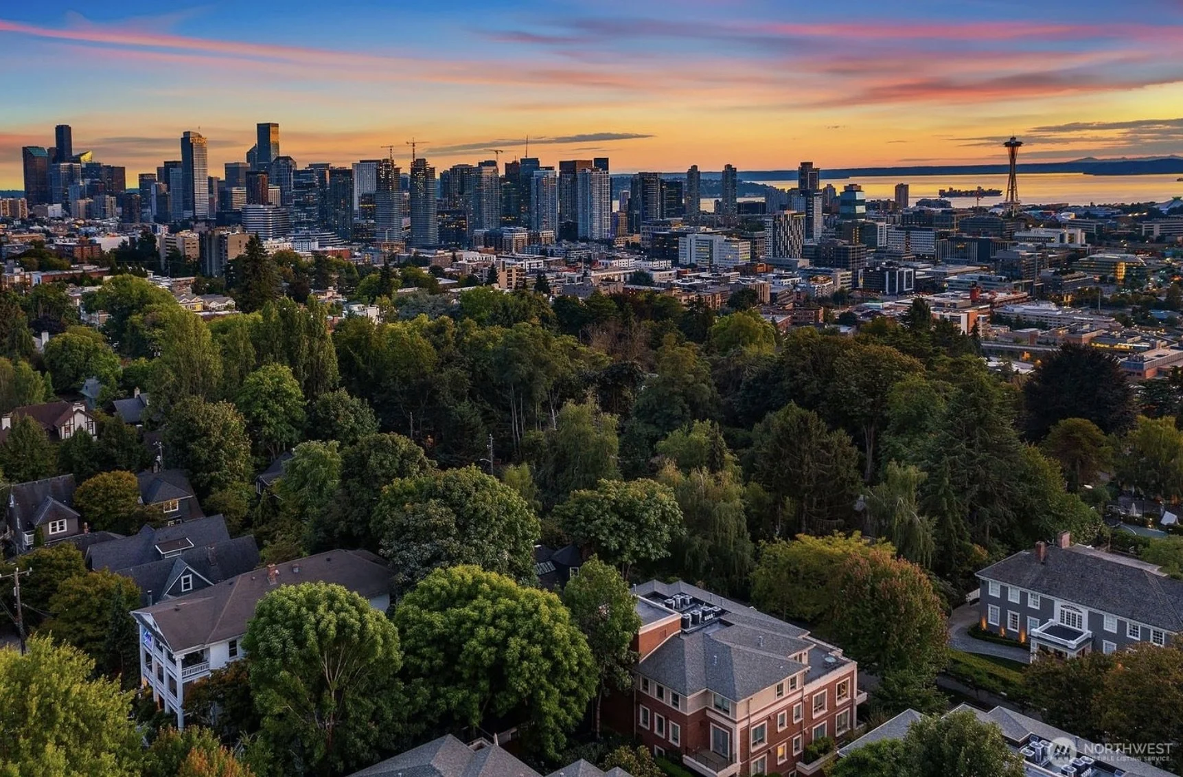 A city skyline at sunset viewed from a hill with residential houses and trees in the foreground.