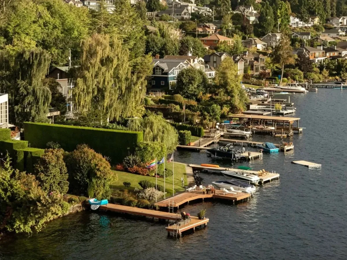 Aerial view of a hillside residential neighborhood near a large body of water, with houses, trees, and a partly cloudy sky.