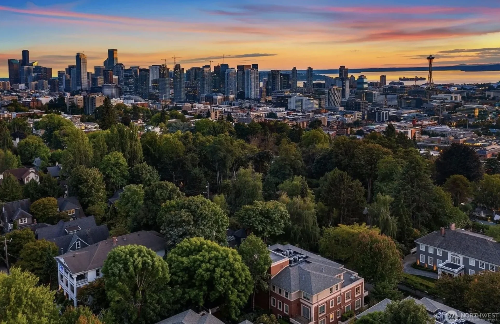 A city skyline at sunset viewed from a hill with residential houses and trees in the foreground.