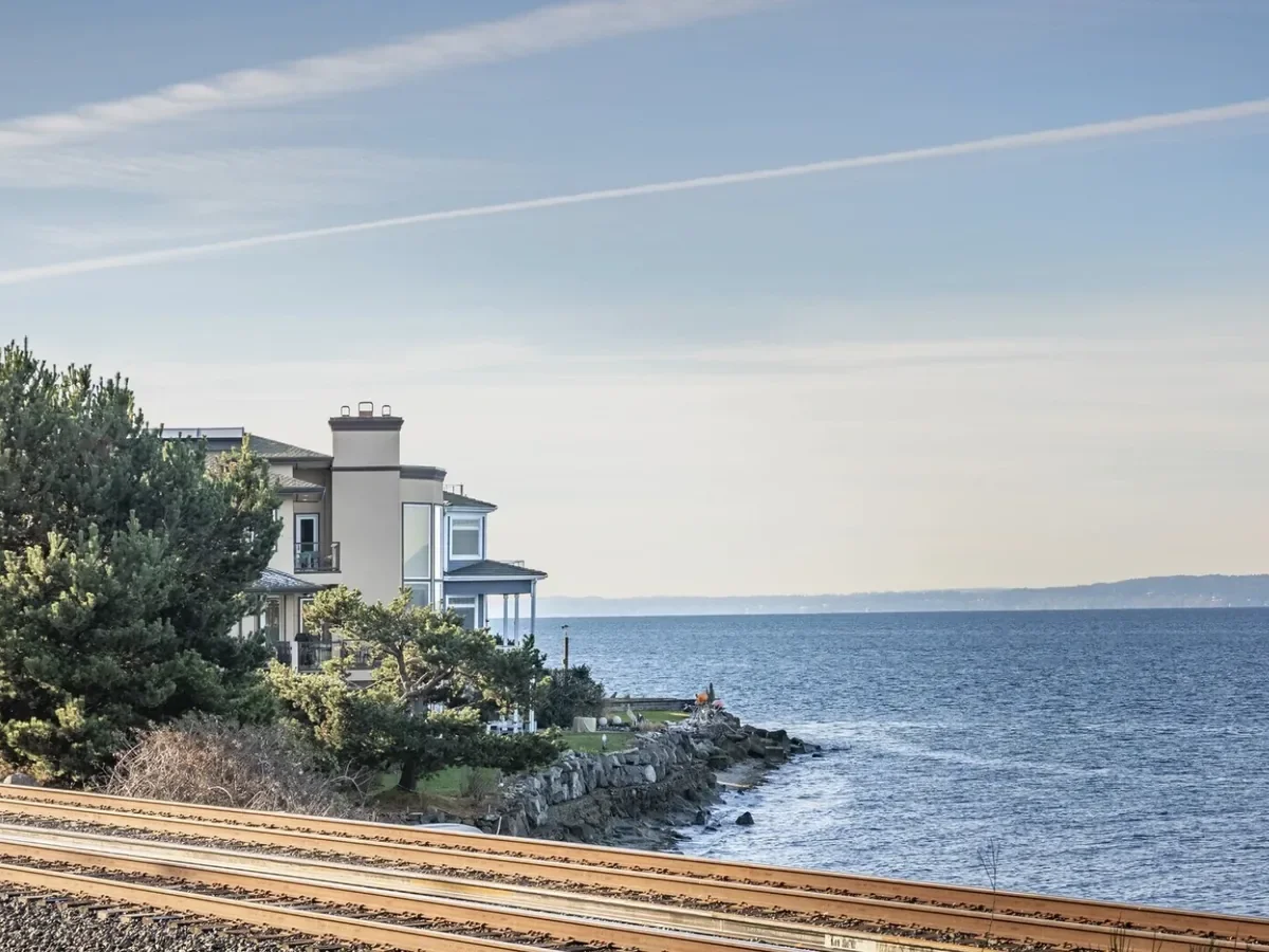 A coastal scene with train tracks in the foreground, a house with trees near the water, and a large body of water extending to the horizon under a partly cloudy sky.