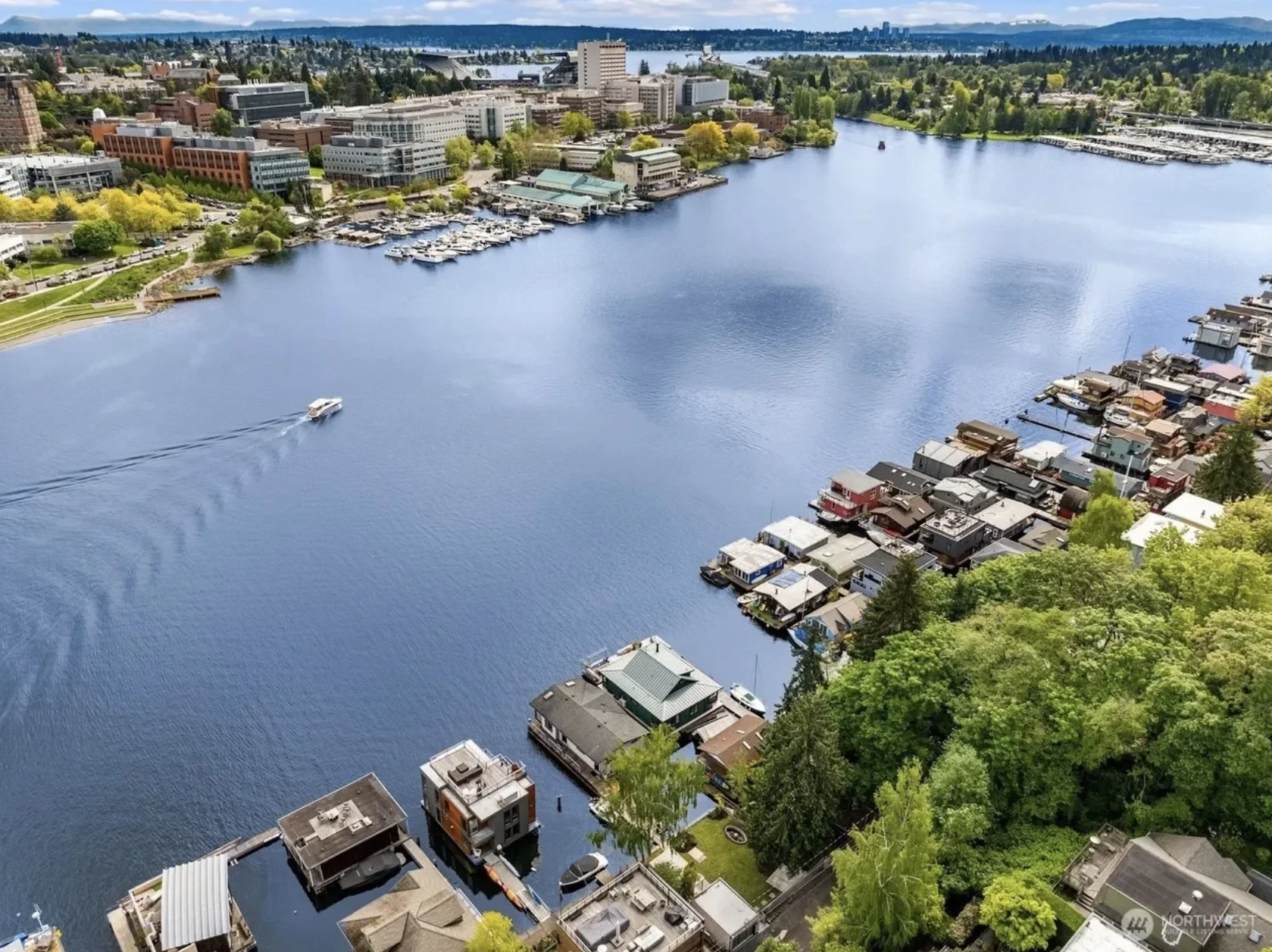 Aerial view of a city by the water featuring a boat on the river, residential houses along the shoreline, and buildings and trees across the river in the background.
