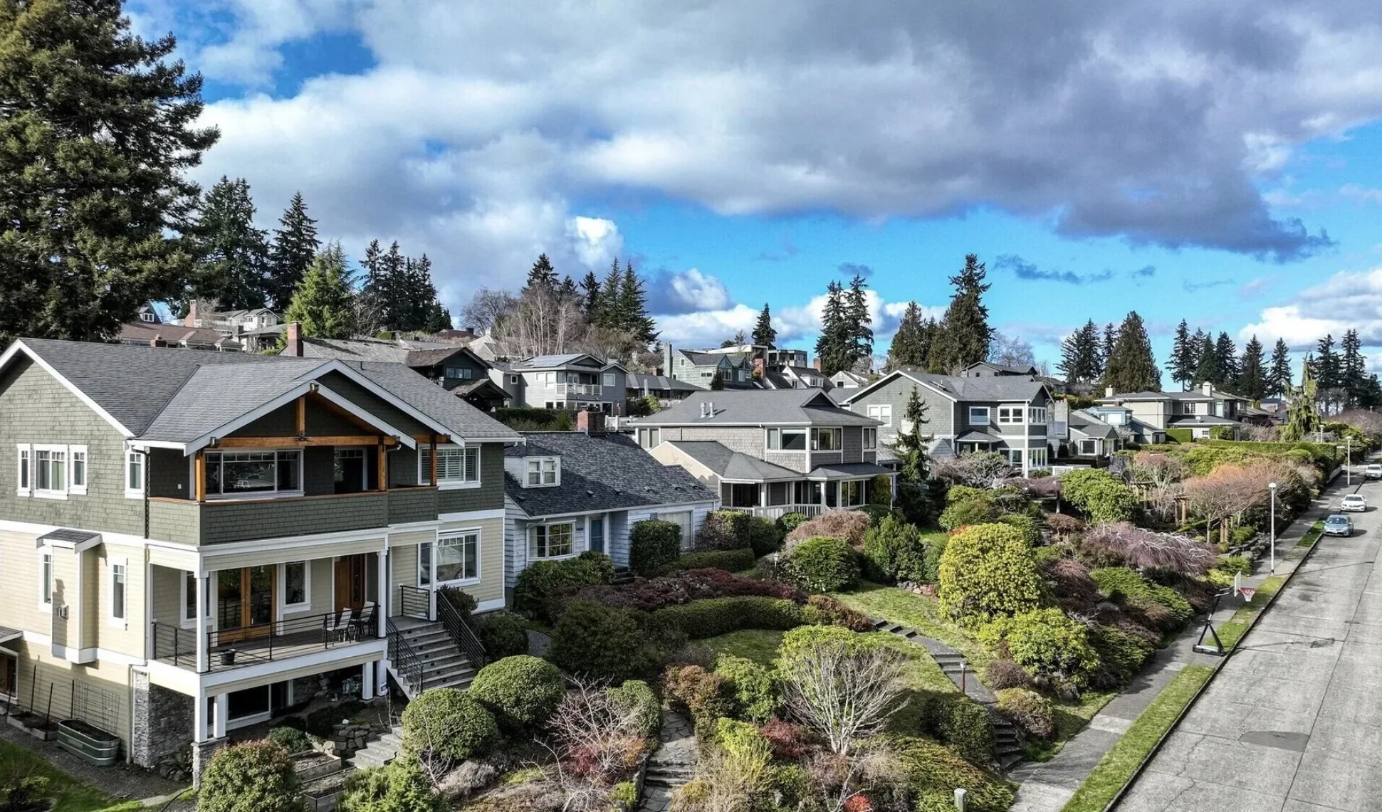 A neighborhood of multi-story houses with well-maintained gardens, trees, and shrubs on a hilly street under a blue sky with clouds.