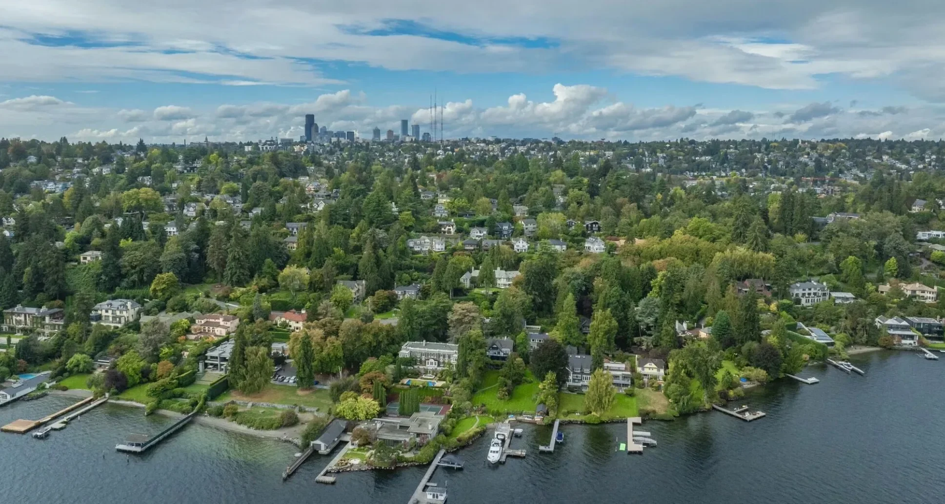 Aerial view of a lakeside residential neighborhood with numerous houses, docks, and boats along the waterfront, surrounded by lush green trees and a distant city skyline under a partly cloudy sky.