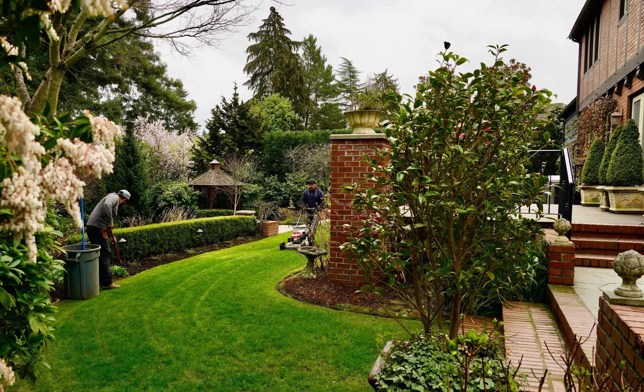 Two men gardening in a lush backyard with green grass, trees, and a gazebo in the background.