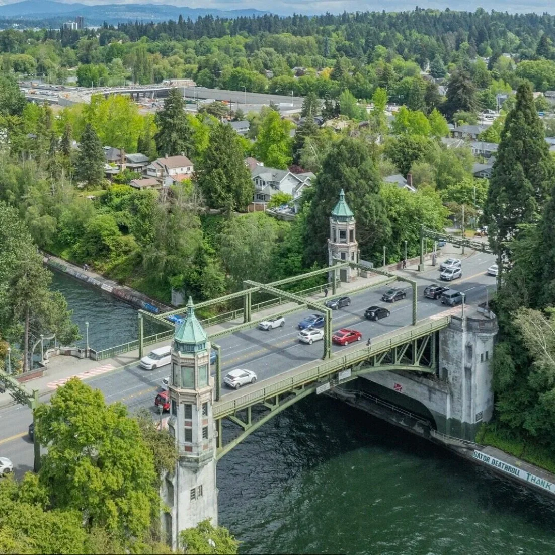 An aerial view of a bridge crossing a river, surrounded by lush green trees and a residential neighborhood with houses and roads in the background.