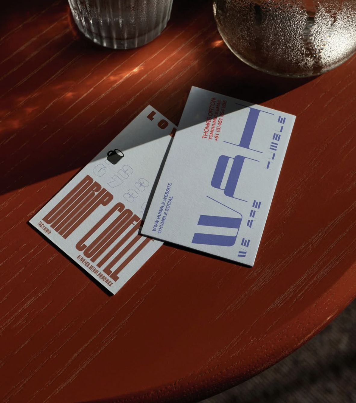 A wooden table with two business cards and two glass cups. One card is from a coffee shop, and the other contains information for a tattoo studio.
