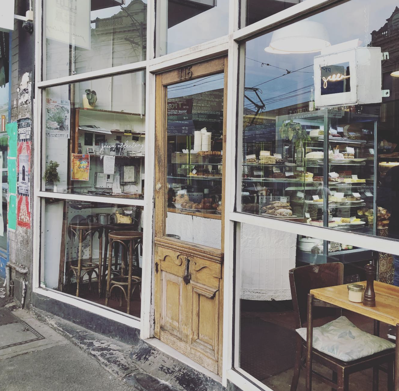 View of a bakery storefront with large glass windows displaying baked goods inside. There is a wooden door and vintage-style chairs visible through the window, with reflections of buildings and power lines outside.