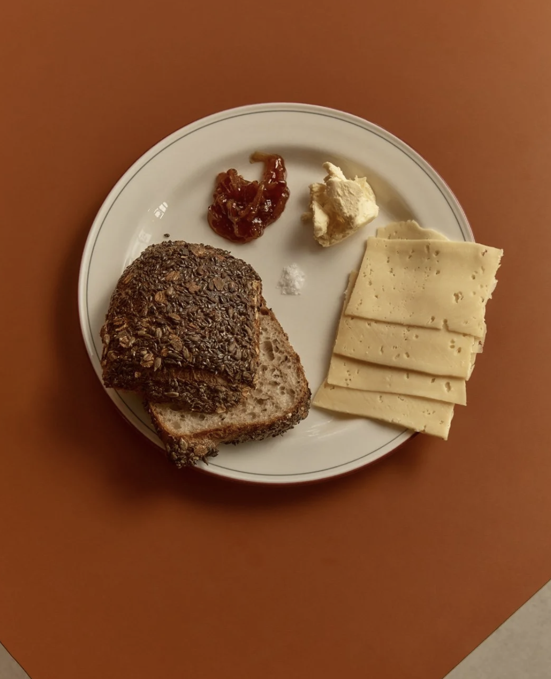 A white plate with slices of Swiss cheese, a piece of bread with seeds, and small portions of caramelized onions and butter on a brown table.