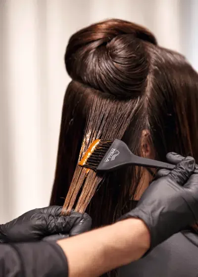 A hairdresser applies hair dye to a client's hair using a brush and gloves.