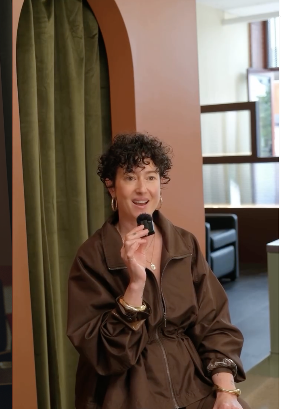 A woman with curly short hair holding a microphone, wearing a brown jacket and jewelry, standing in a room with green curtains and large windows.