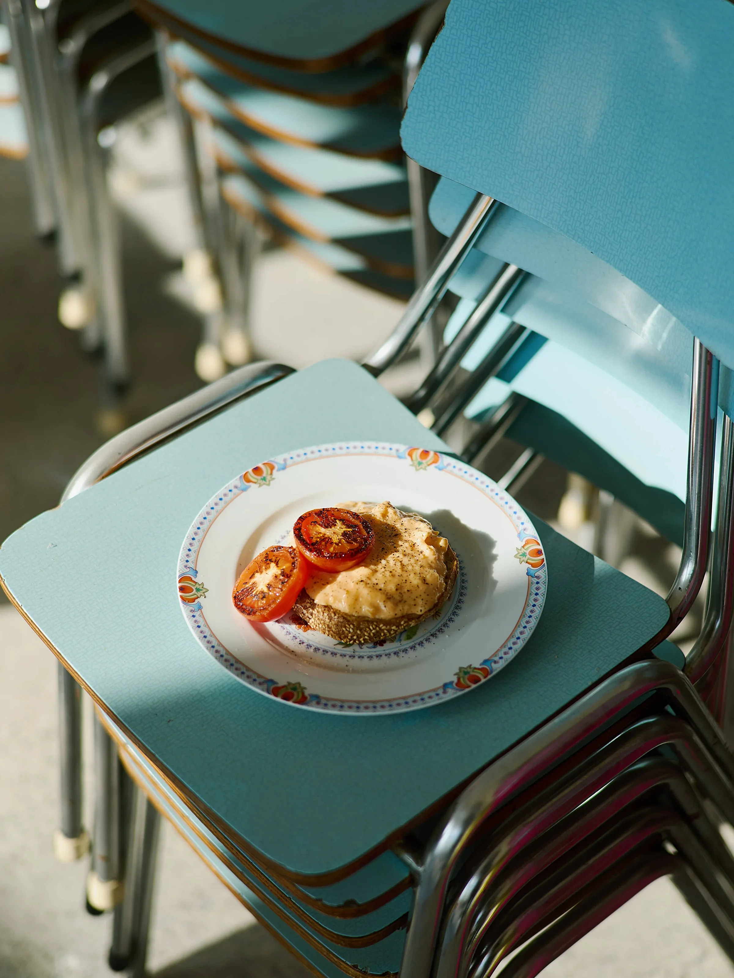 A plate of food with sliced tomatoes on top, resting on a teal table with a stack of chairs in the background.