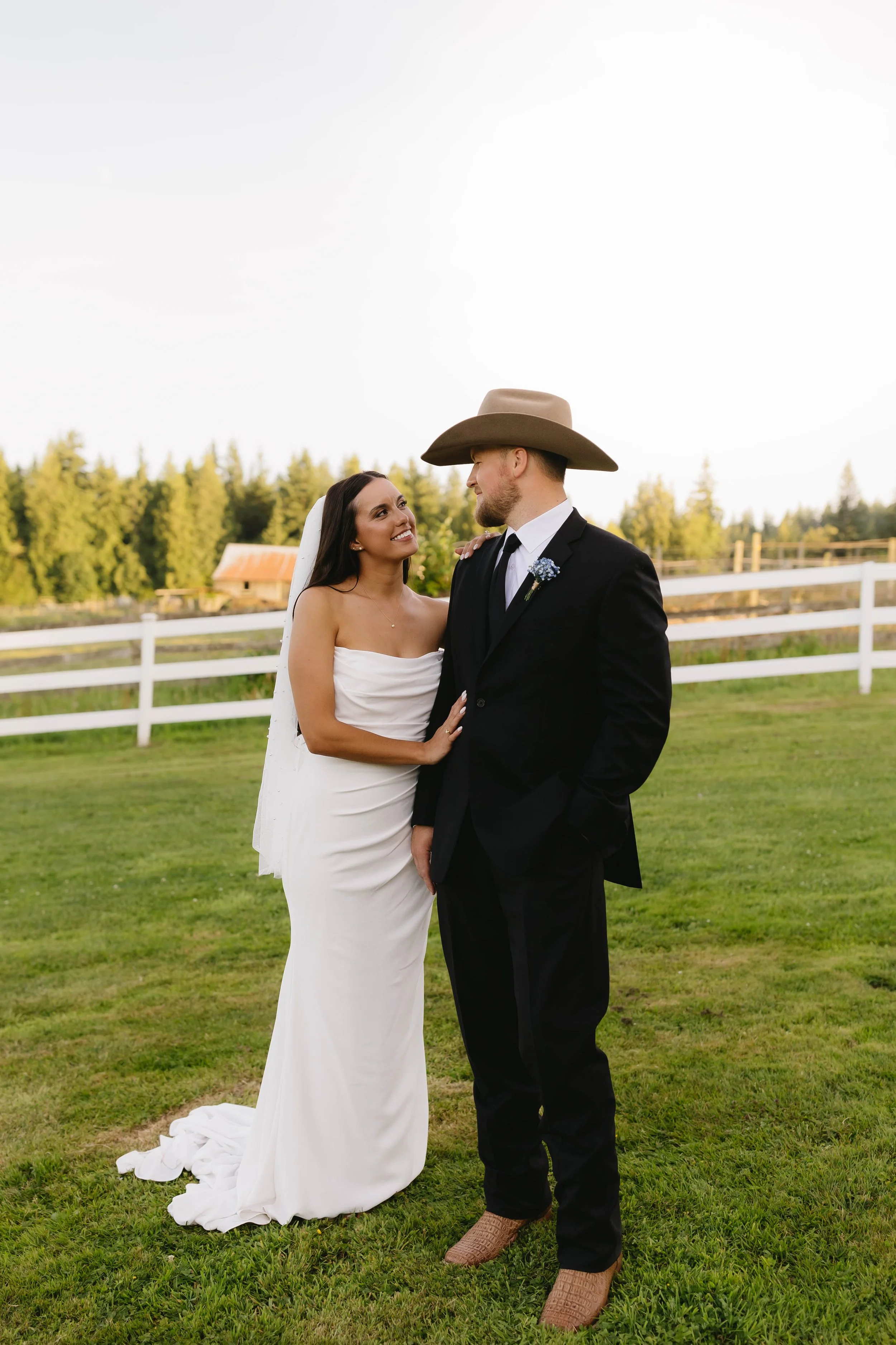A bride and groom standing on green grass outdoors during sunset, gazing at each other. The bride is in a strapless white wedding gown with a veil, and the groom is in a black suit with a white shirt, a tie, a boutonnière, and a cowboy hat. There are