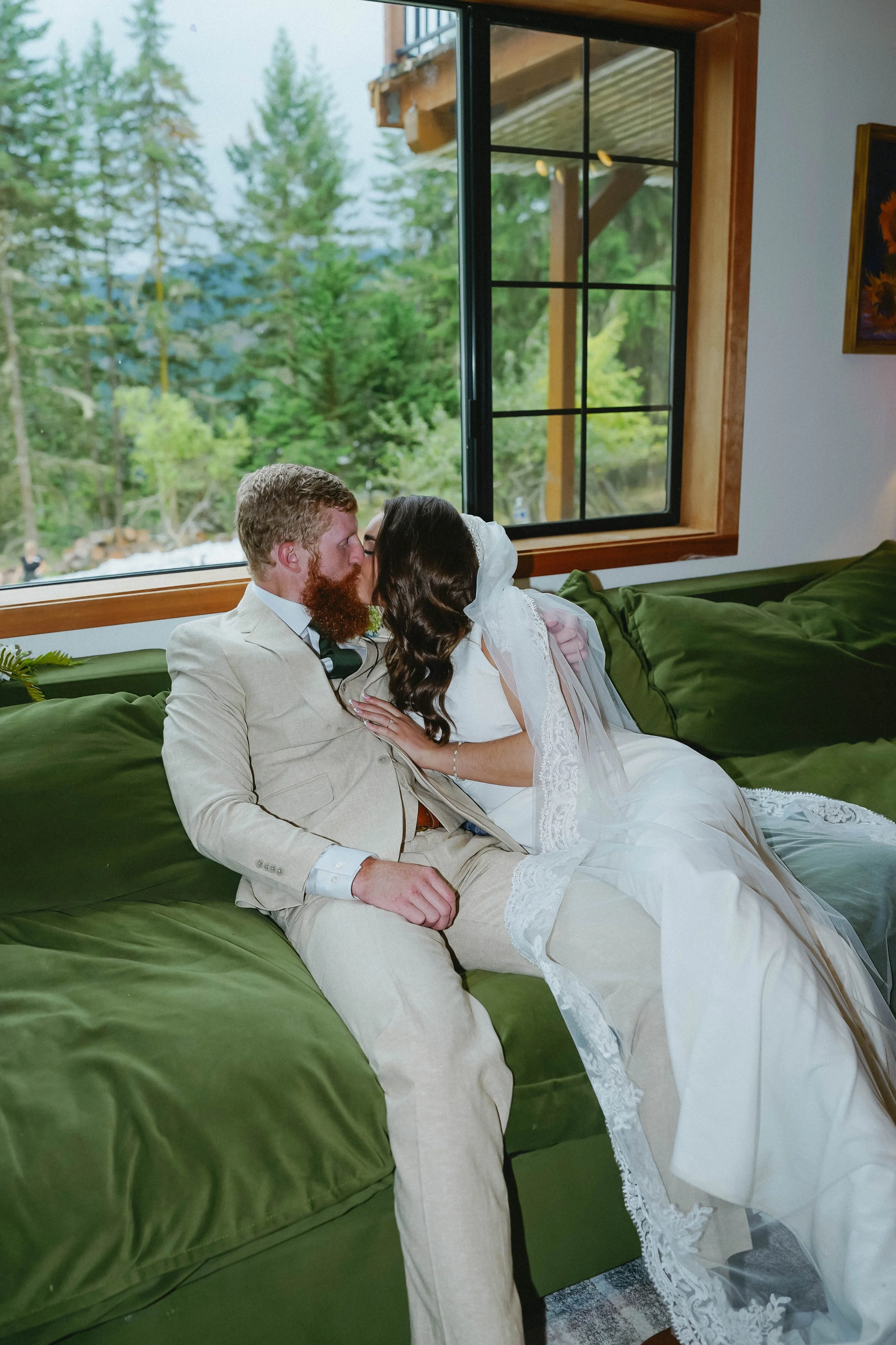 A bride and groom sit on a green couch inside, sharing a kiss in front of a large window showing trees outside.