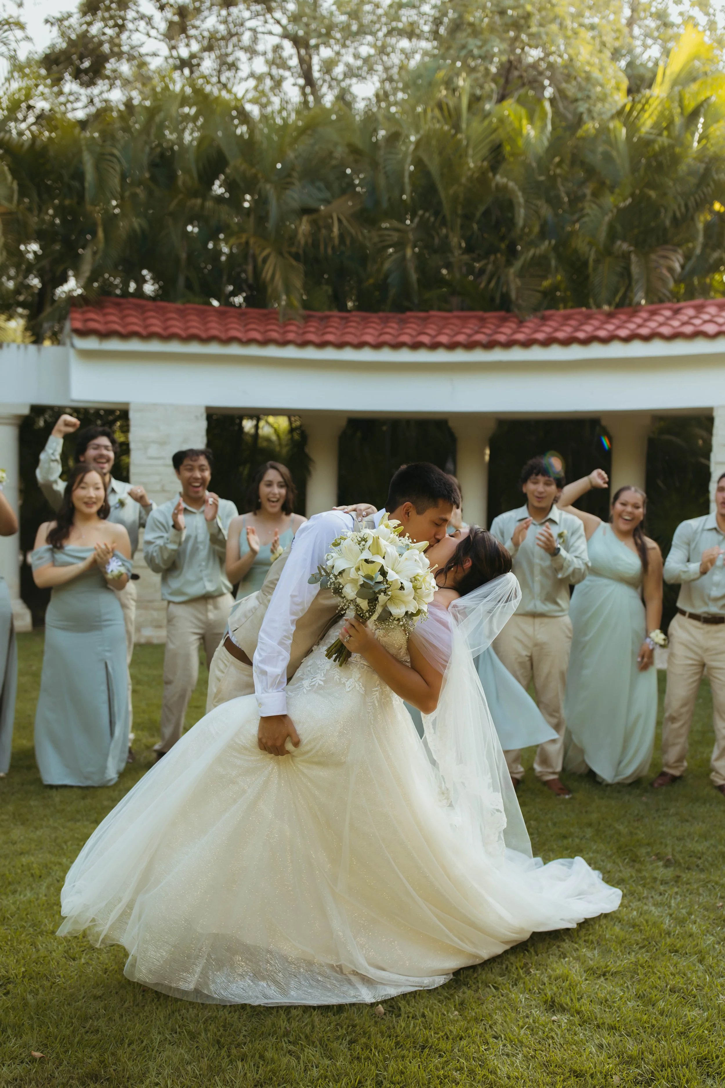 A bride and groom share a kiss during their wedding ceremony outdoors, surrounded by friends and family, with lush trees and a building in the background.