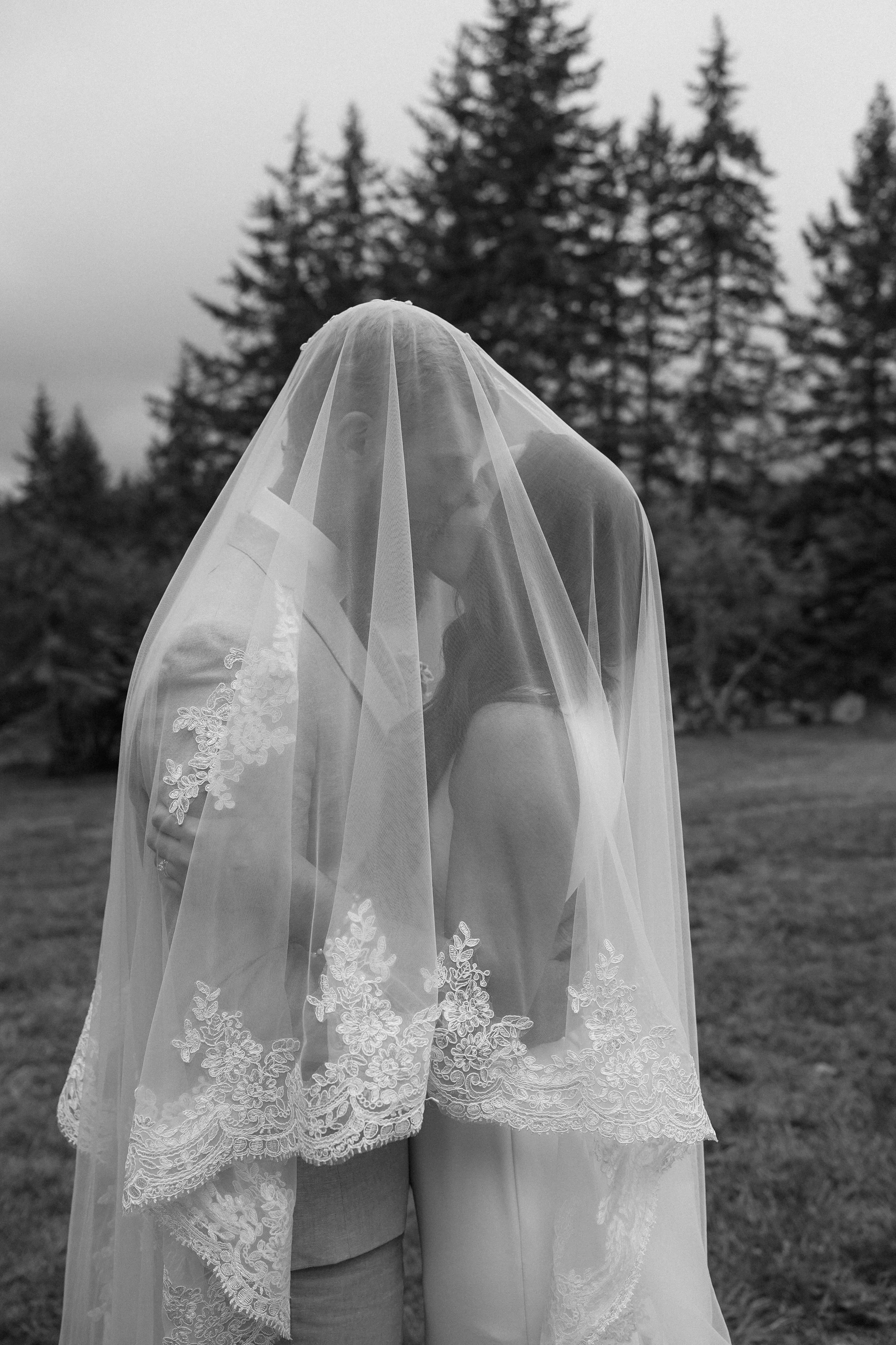 Black and white photo of a bride and groom kissing under a lace veil outdoors with trees in the background.