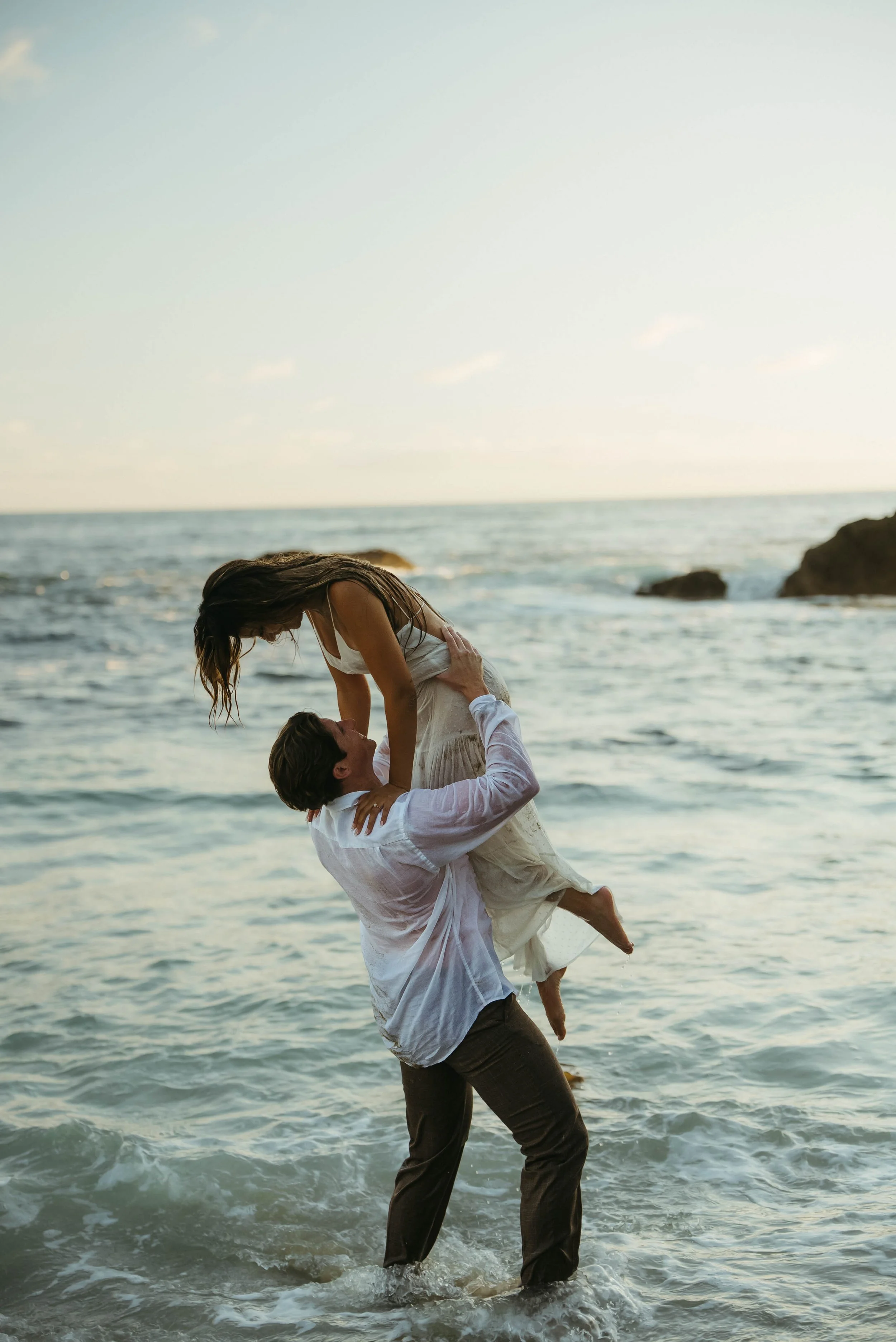 A couple at the beach, with the man lifting the woman in his arms while standing in the water, during sunset or late afternoon.