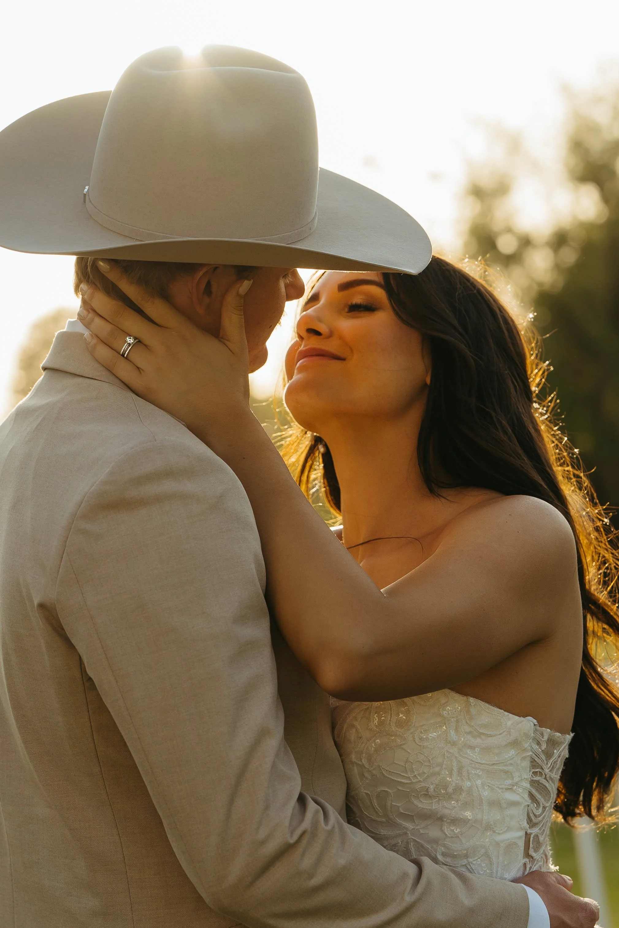 A couple embraces outdoors at sunset, with the woman holding the man's face. The man wears a light-colored suit and cowboy hat, while the woman wears a strapless wedding dress, both smiling softly.