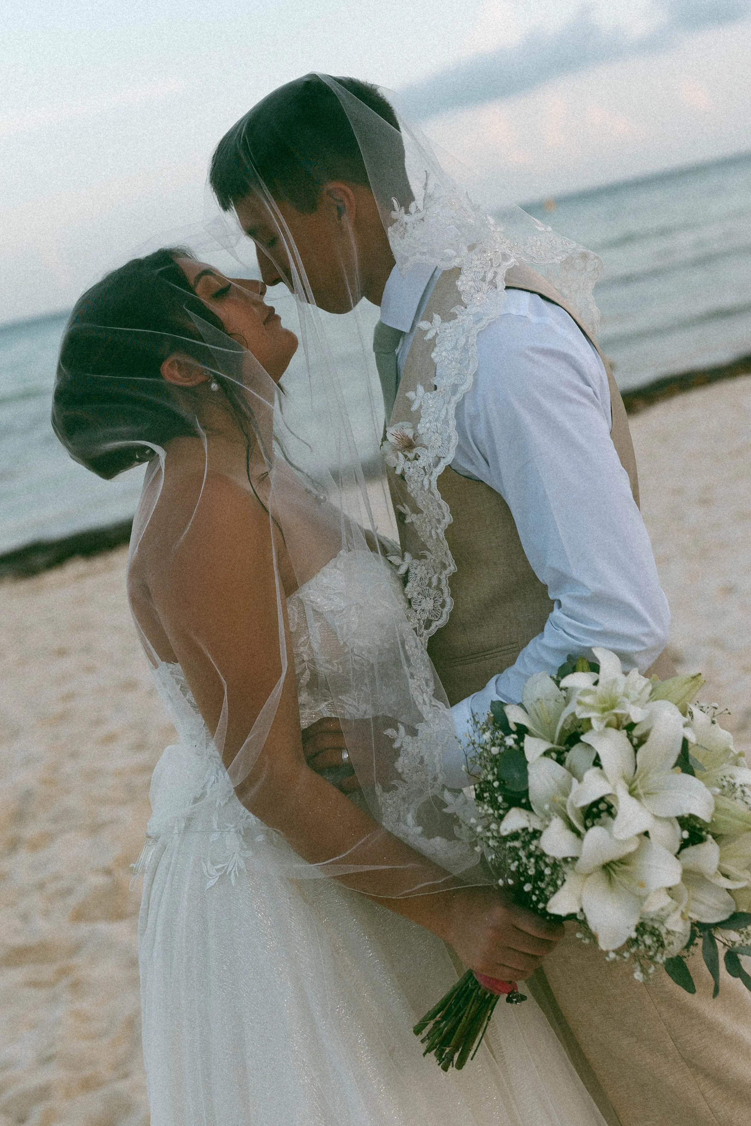 A bride and groom, both with wedding veils, standing close and about to kiss on a beach, holding a bouquet of white lilies and greenery.