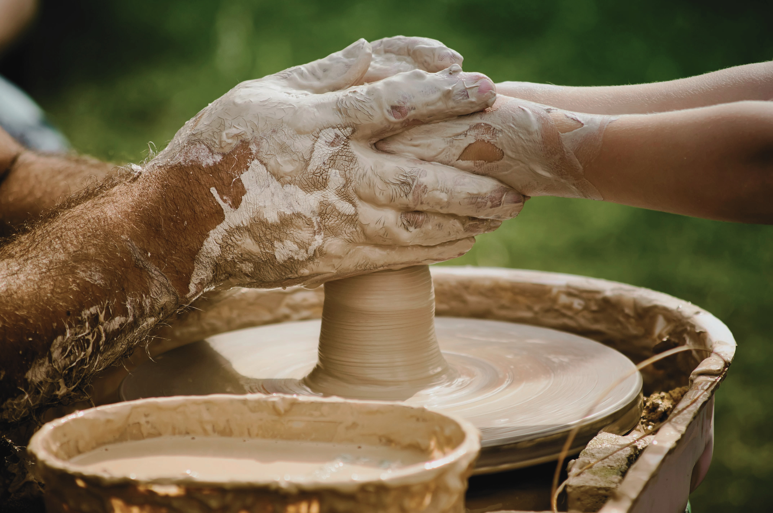 Close-up of two people practicing pottery, with one person guiding the other's hands on a spinning clay wheel.