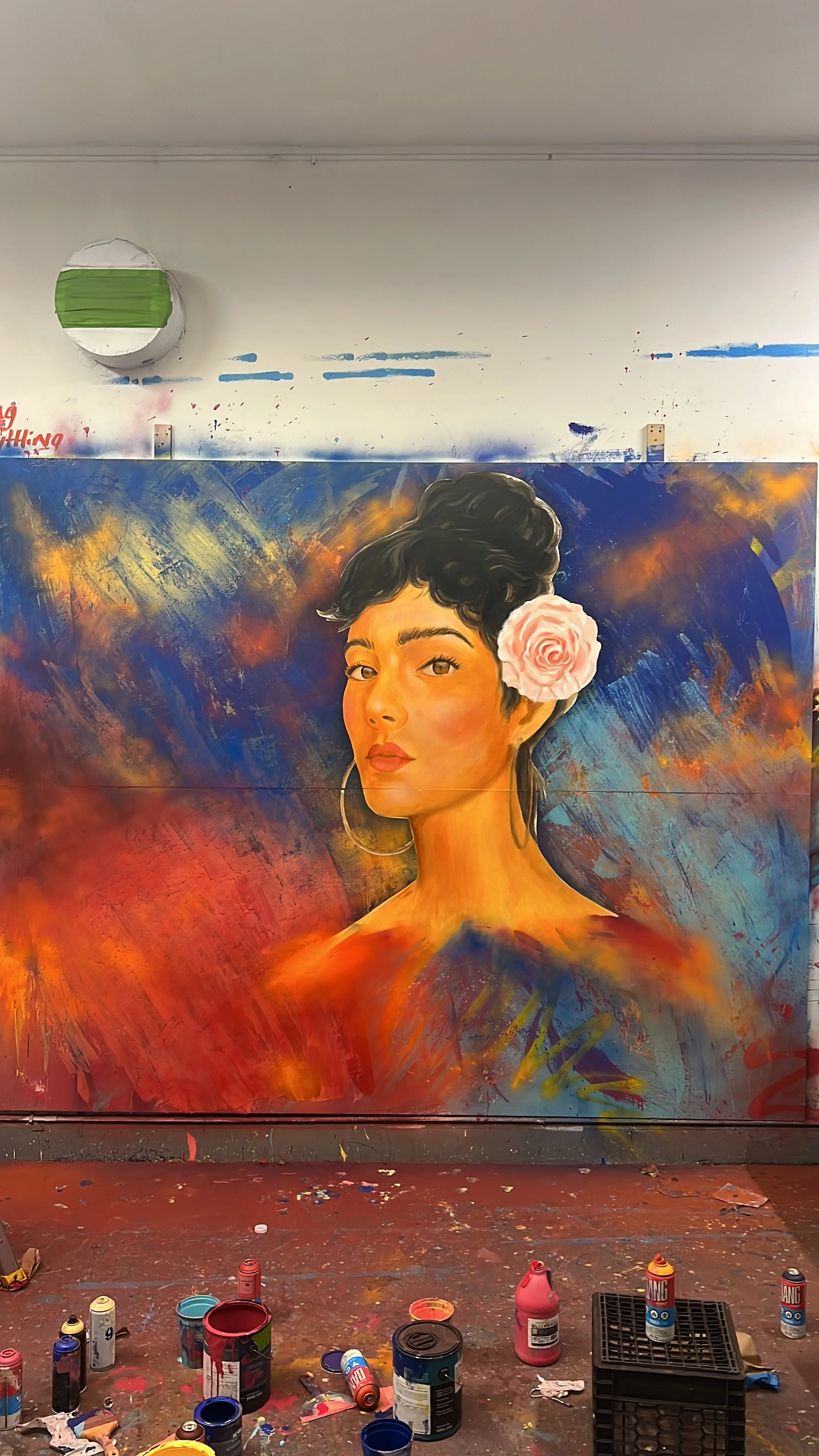A colorful portrait of a woman with dark hair styled in an updo, adorned with a pink flower, painted on a large canvas surrounded by paint cans and splatters in an art studio.