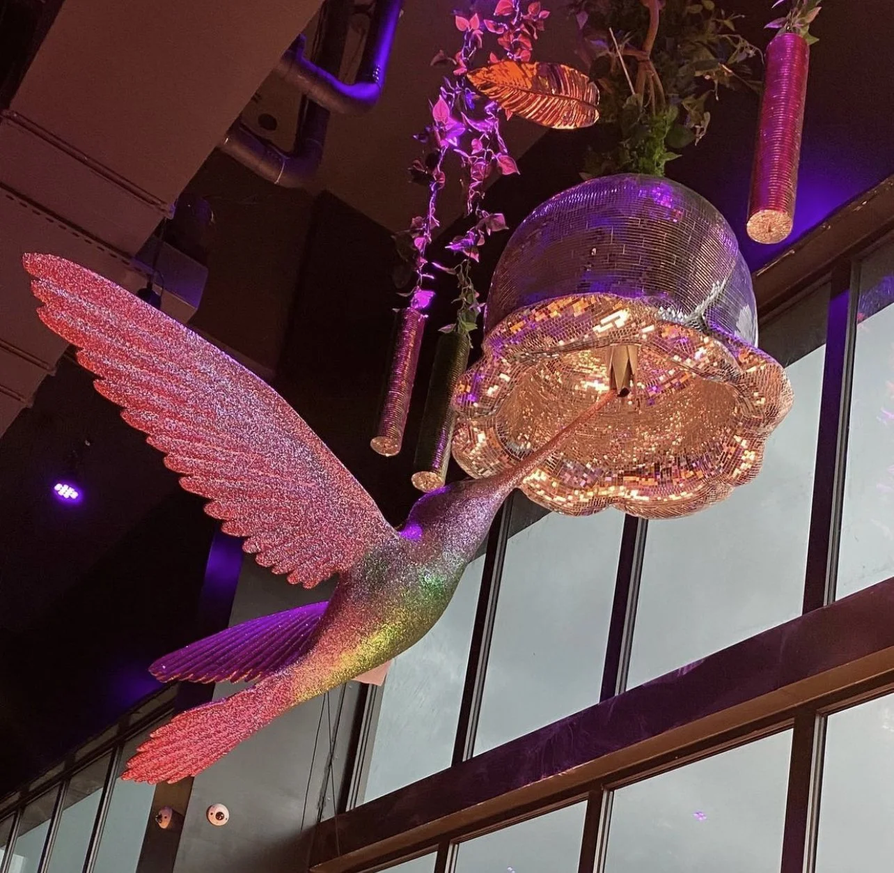 A decorative sculpture of a hummingbird with pink, green, and purple glittering details, positioned in front of a glass window. The hummingbird's beak is extended toward a large, illuminated, flower-shaped art piece made of small reflective tiles, su