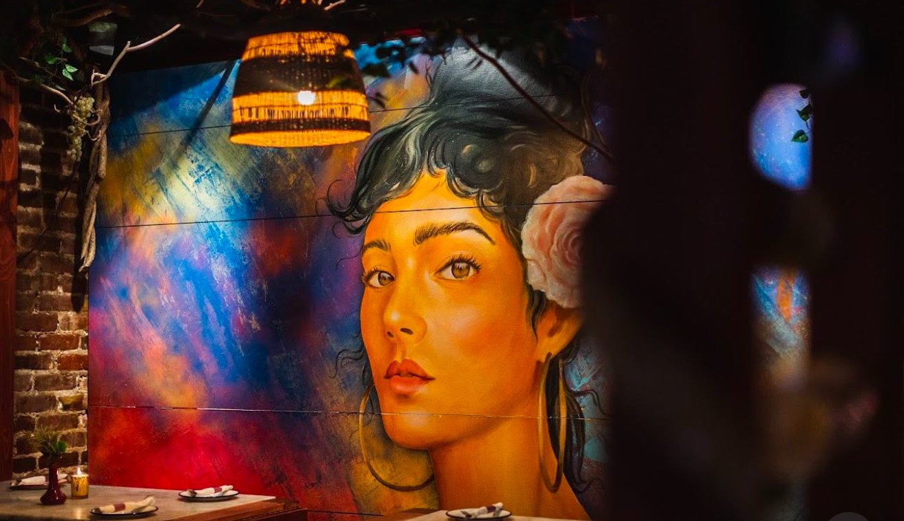 Colorful mural of a woman's face on a wall in a restaurant with a hanging lamp and table setting in foreground.