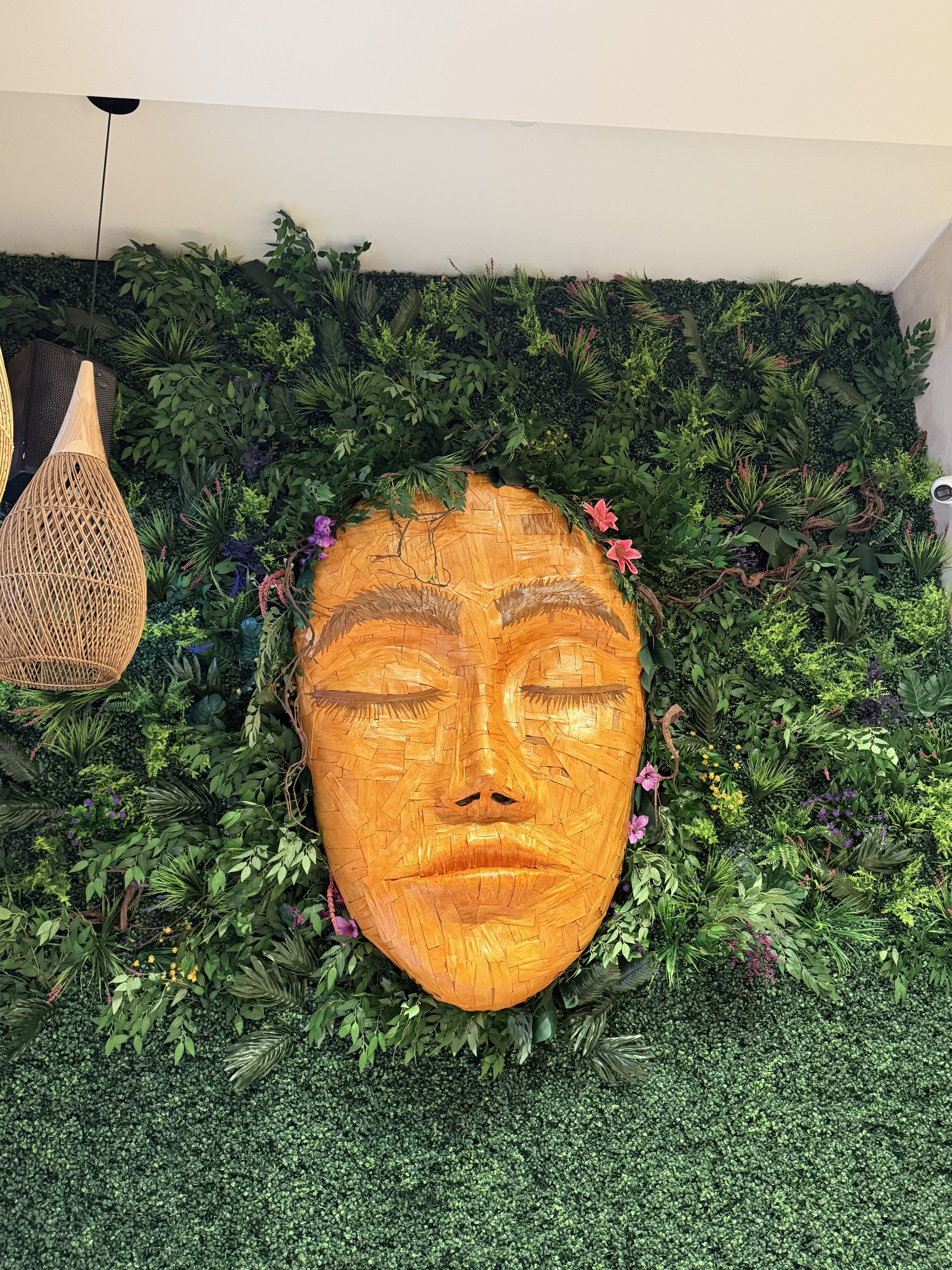 A large wall art sculpture portrait of a face with closed eyes, surrounded by lush green plants and flowers. Tulum vibe background