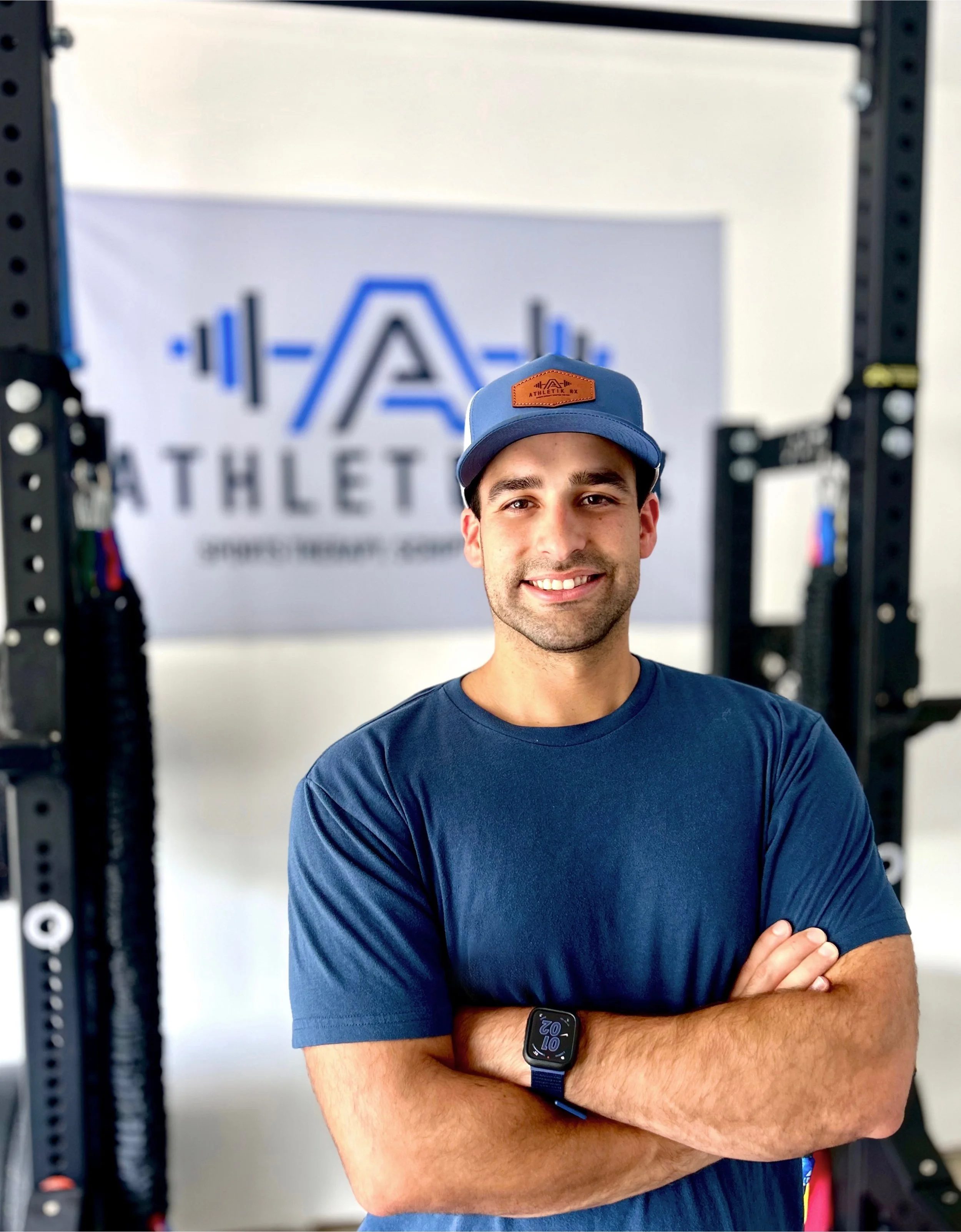 A young man wearing a blue t-shirt, a blue cap with a brown patch, and a smartwatch on his wrist, standing with arms crossed in front of a gym or fitness center backdrop. The background features a logo and the word 'ATHLET'.