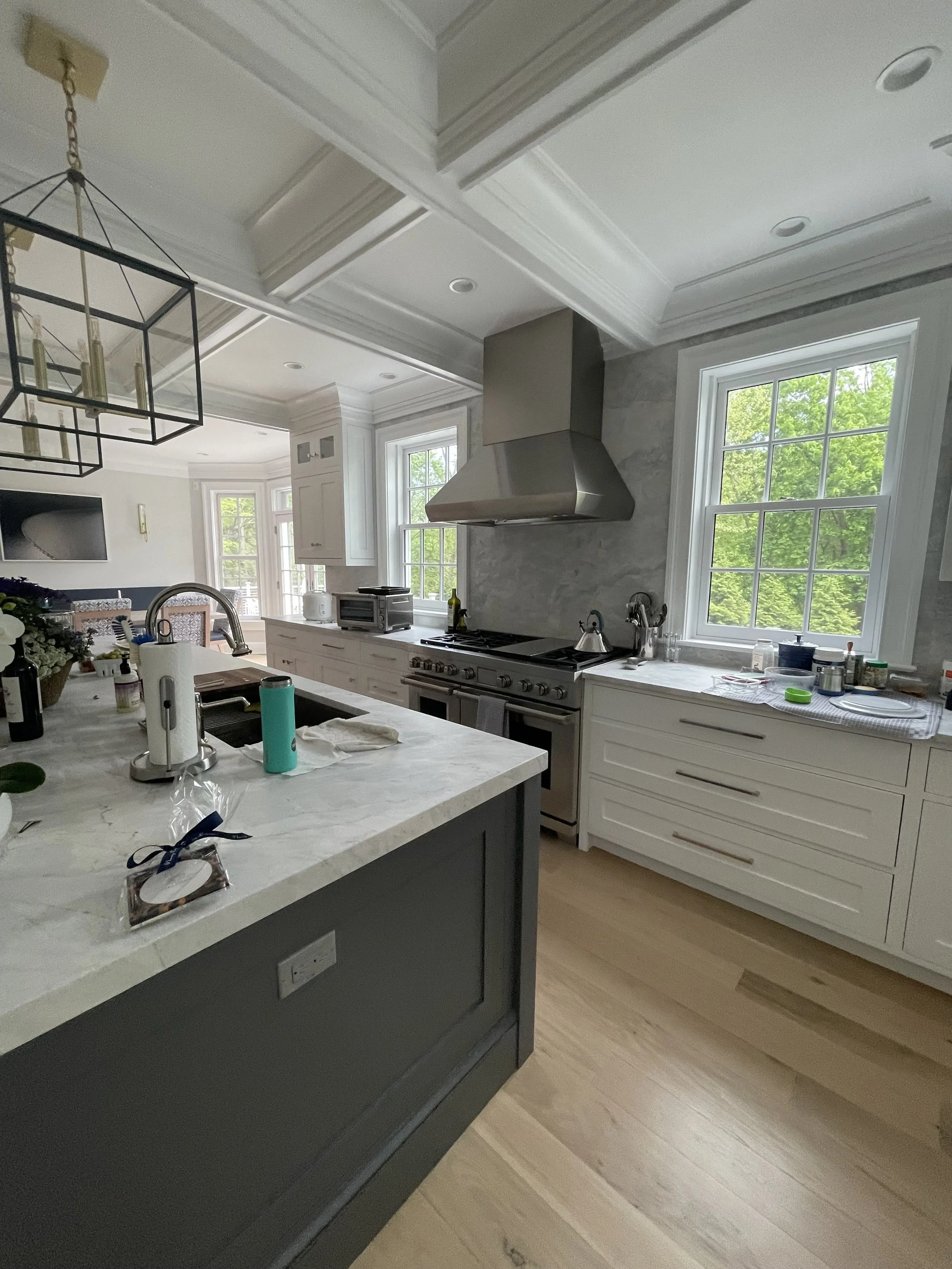 Modern kitchen with white cabinets, stainless steel range hood, marble countertops, and large windows with green trees outside.