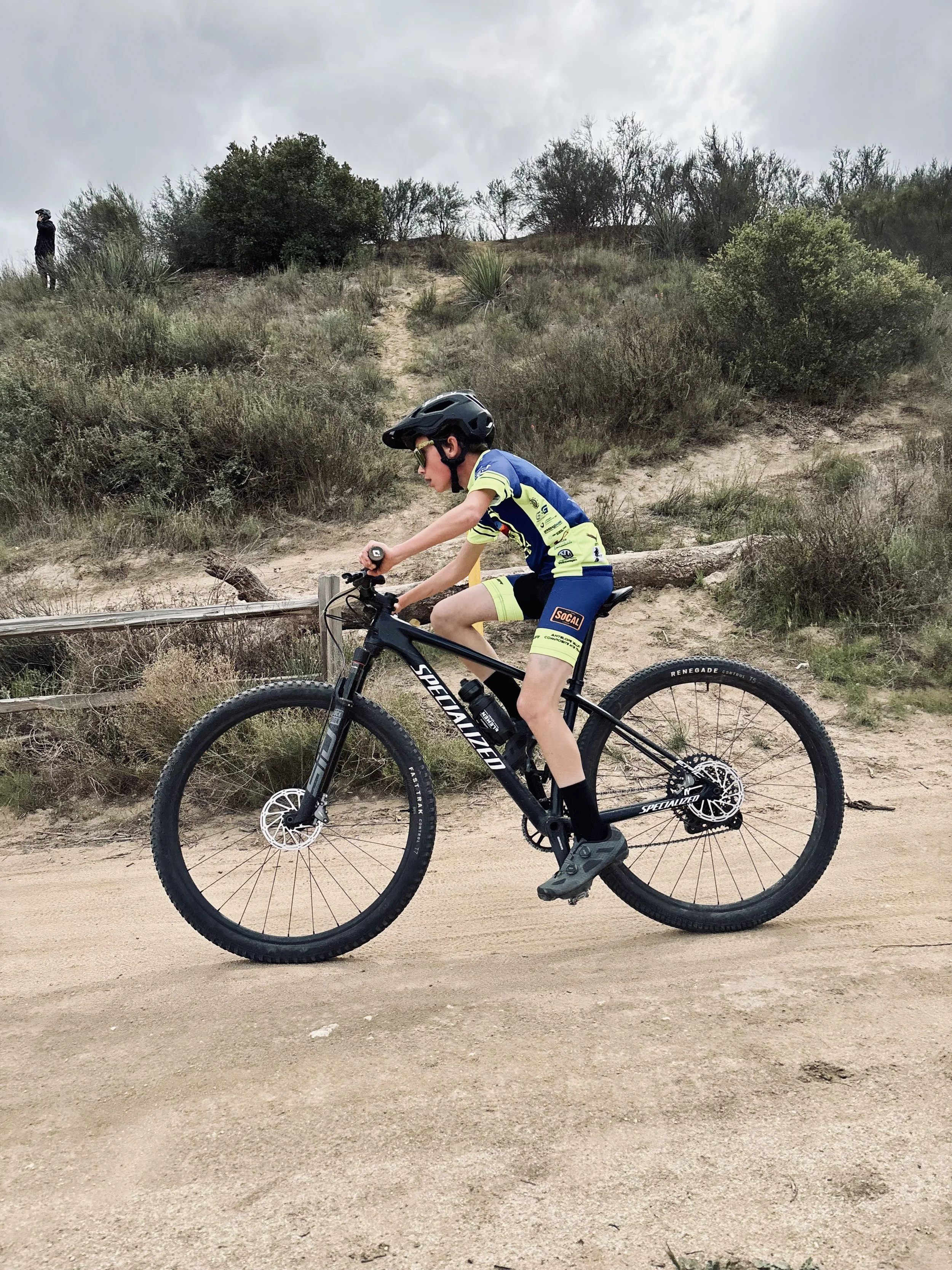 A young boy riding a black mountain bike on a dirt trail in a hilly, shrub-filled area during overcast weather.