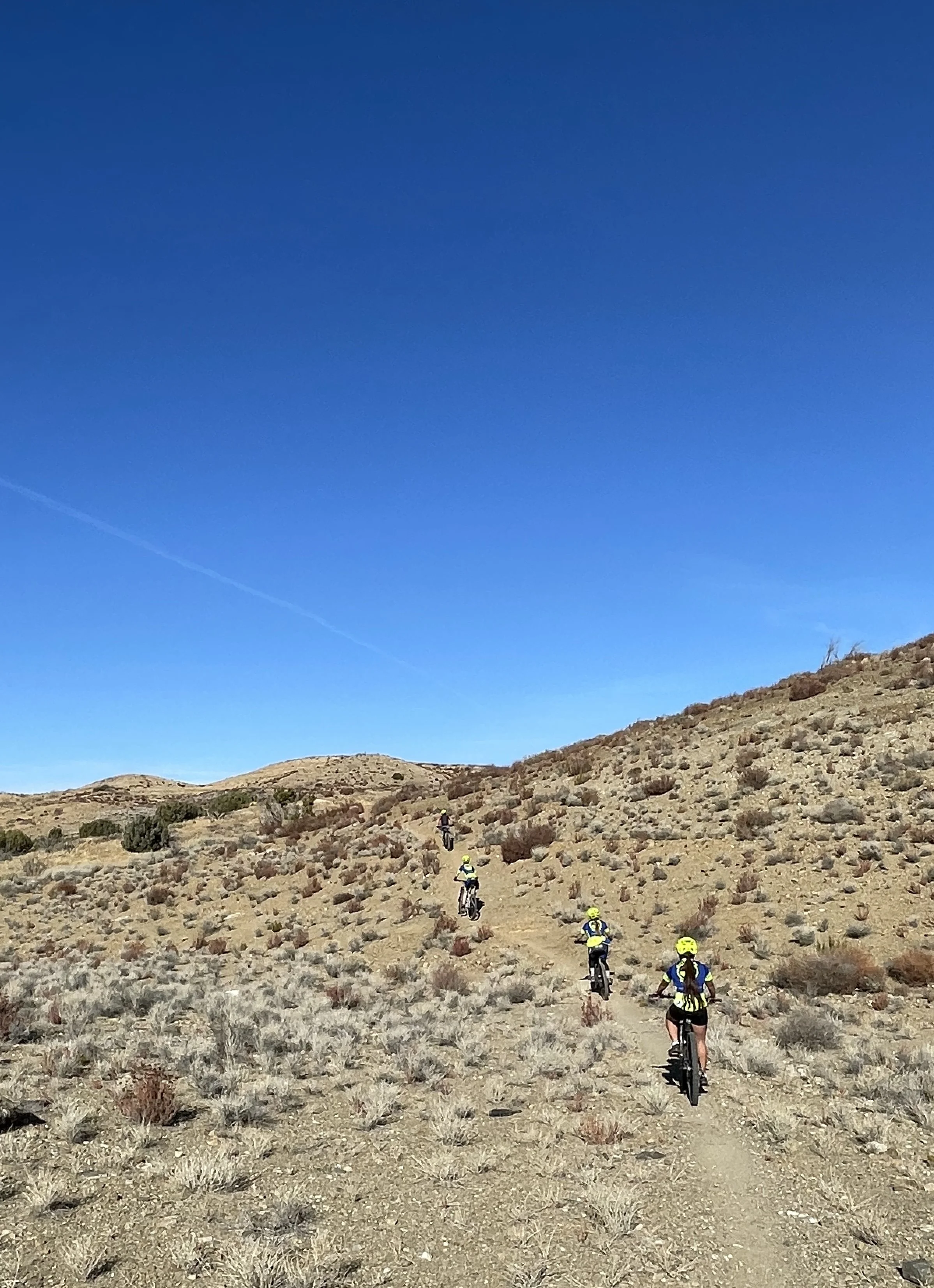 Group of mountain bikers riding on a dirt trail through a dry, rocky desert landscape under a clear blue sky.