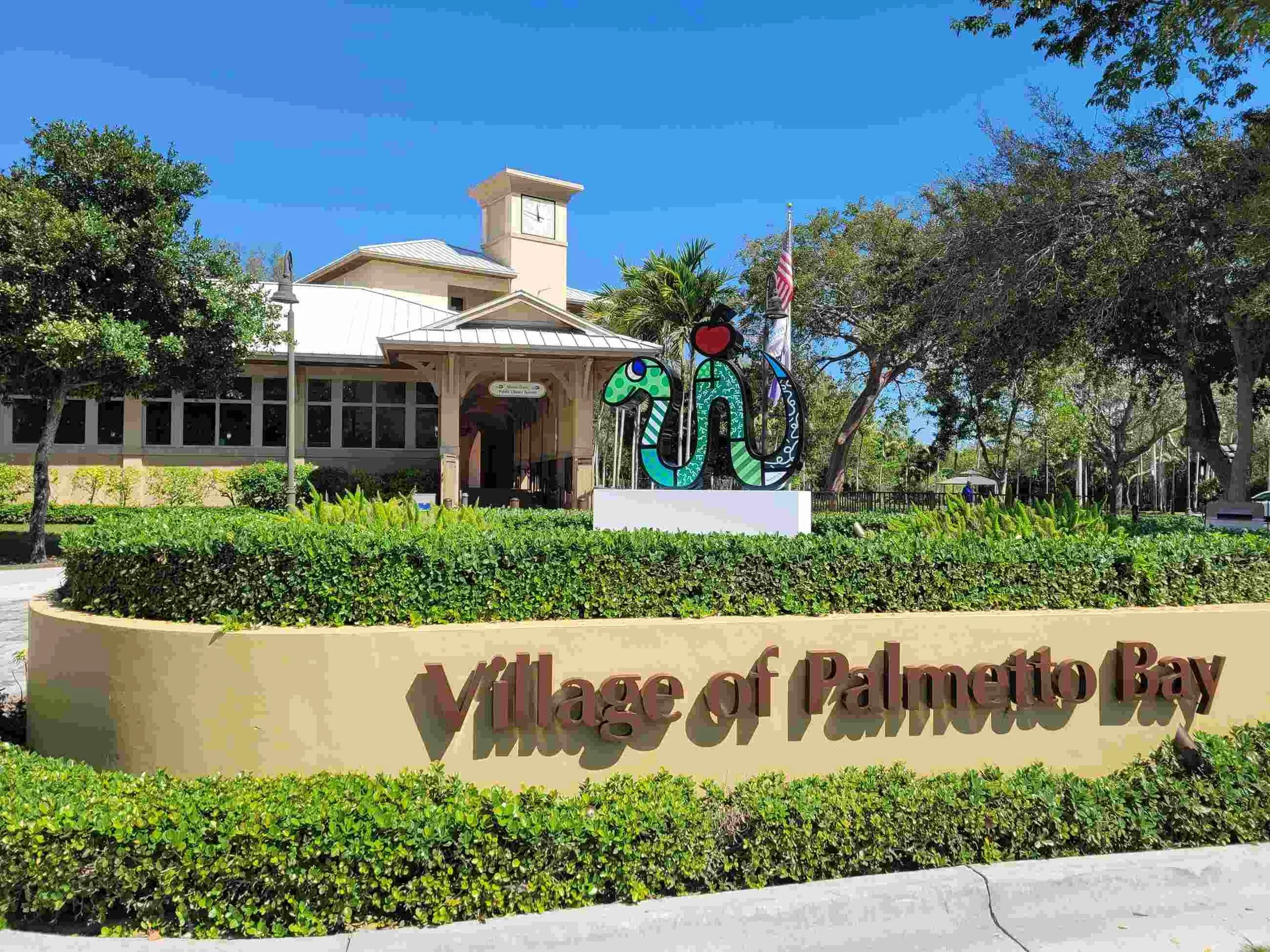 Sign that reads 'village of Palmetto Bay' in front of a building with a clock tower, surrounded by trees, bushes, and decorative art sculptures. Where Mobile IV Nurse provides convenient IV therapy and hydration.