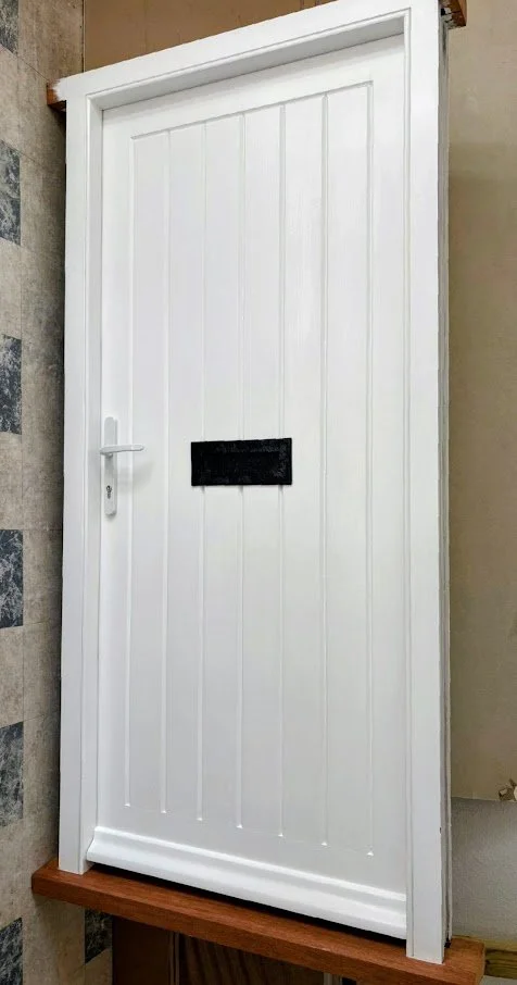 New white interior door with vertical paneling, door handle, and a black mail slot, open in a room with beige walls and wooden floor.