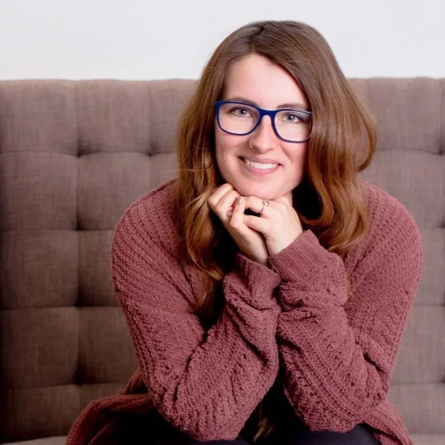 A smiling woman with long red hair and glasses, wearing a maroon sweater, sitting on a beige tufted couch, with hands resting under her chin.