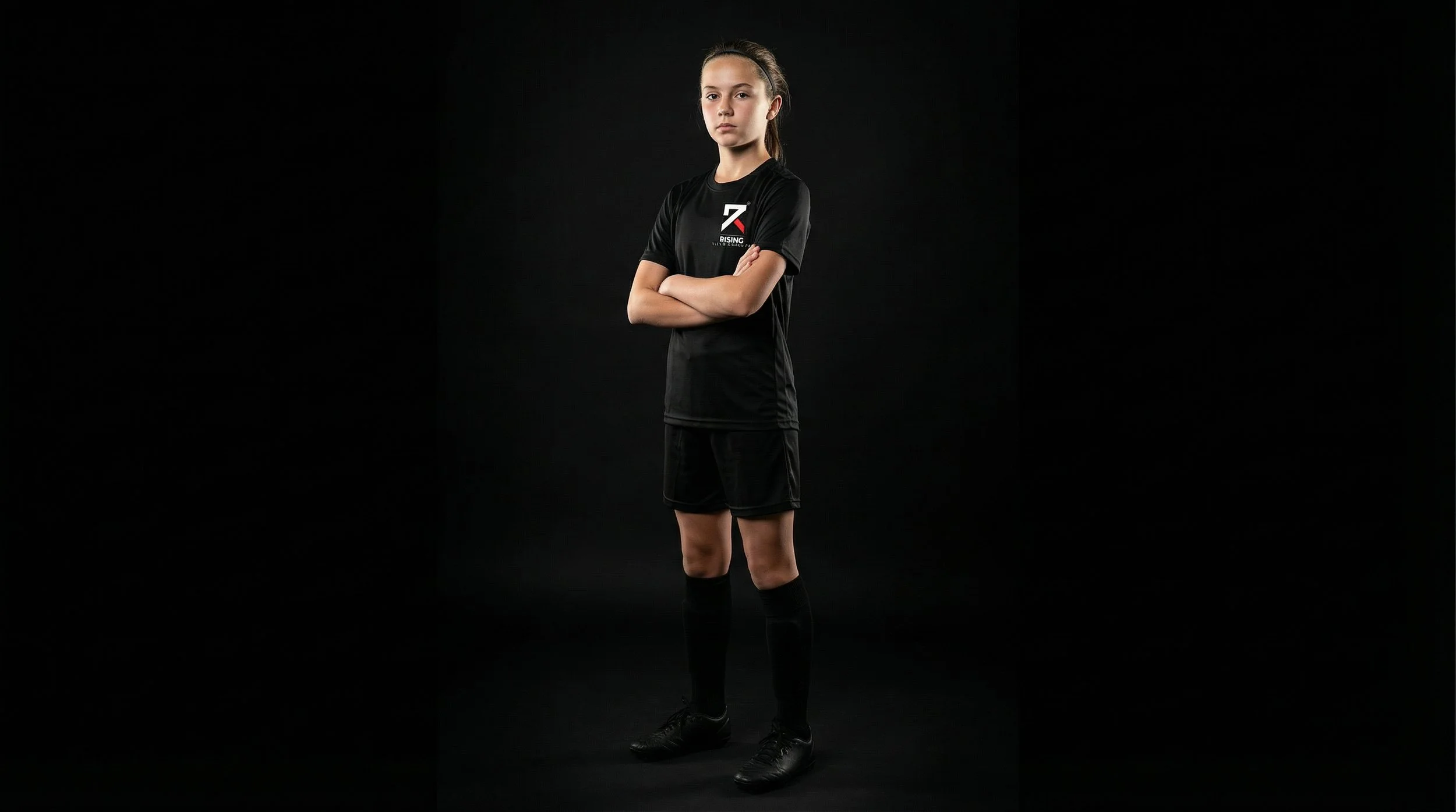 A young female soccer player stands with arms crossed in a black uniform against a black background.