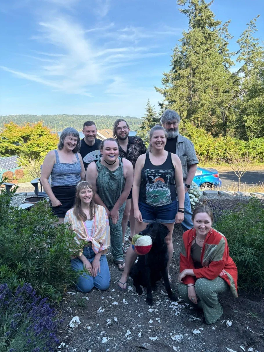 Group of nine people at the 2025 Annex Company retreat, including children and adults, outdoors on a sunny day, crouching and standing around a black dog with a ball in its mouth. Tall trees and a blue car are in the background.