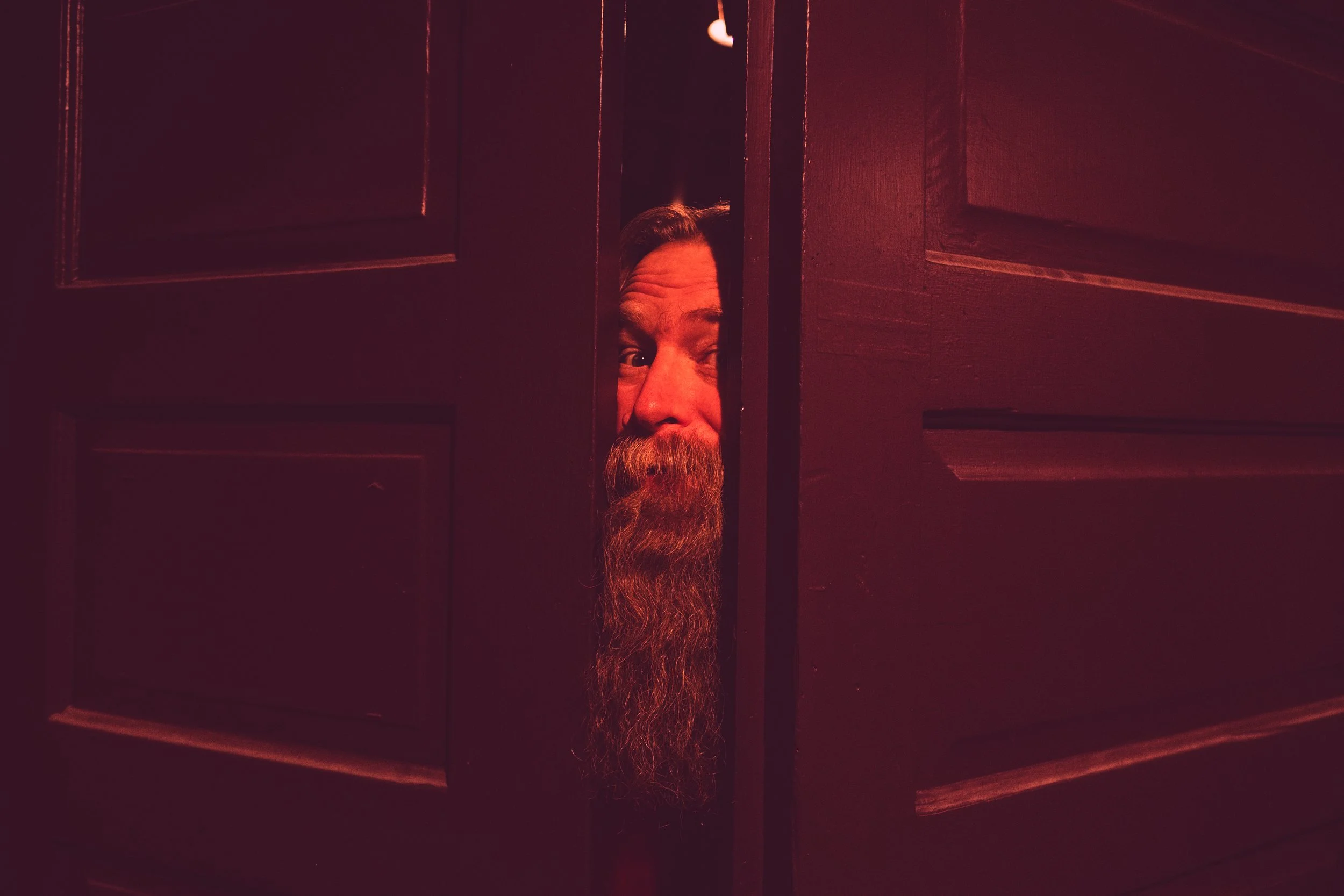 Emmett Montgomery as Sugar Plum Gary, with a long beard peeking through a slightly open dark wooden door, illuminated with red lighting.
