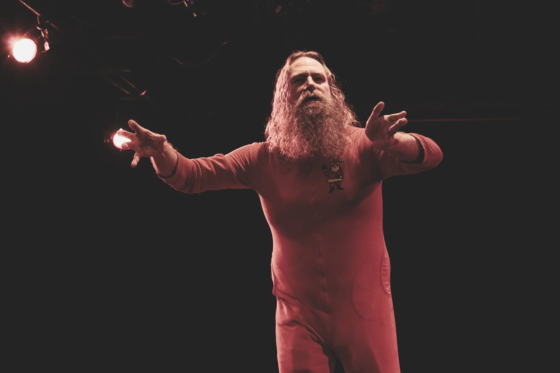 Emmett Montgomery with long hair and a beard performing on stage as Sugar Plum Gary, wearing a pink jumpsuit, with his arms extended and spotlight behind him.