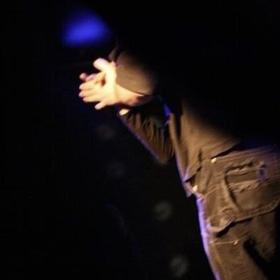 Stephen McCandless on stage with blue spotlight, clapping hands, wearing brown jacket and pants, dark background.