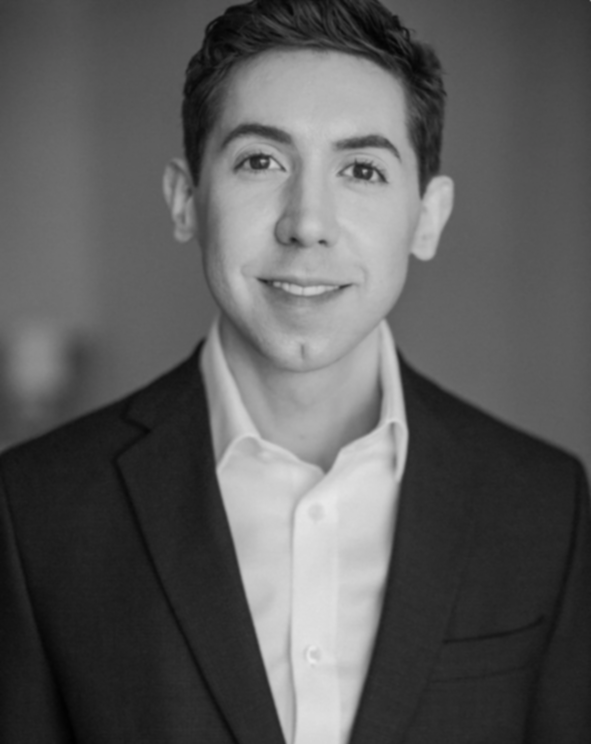 Professional portrait of a young man wearing a suit and white shirt, smiling, with a blurred background.