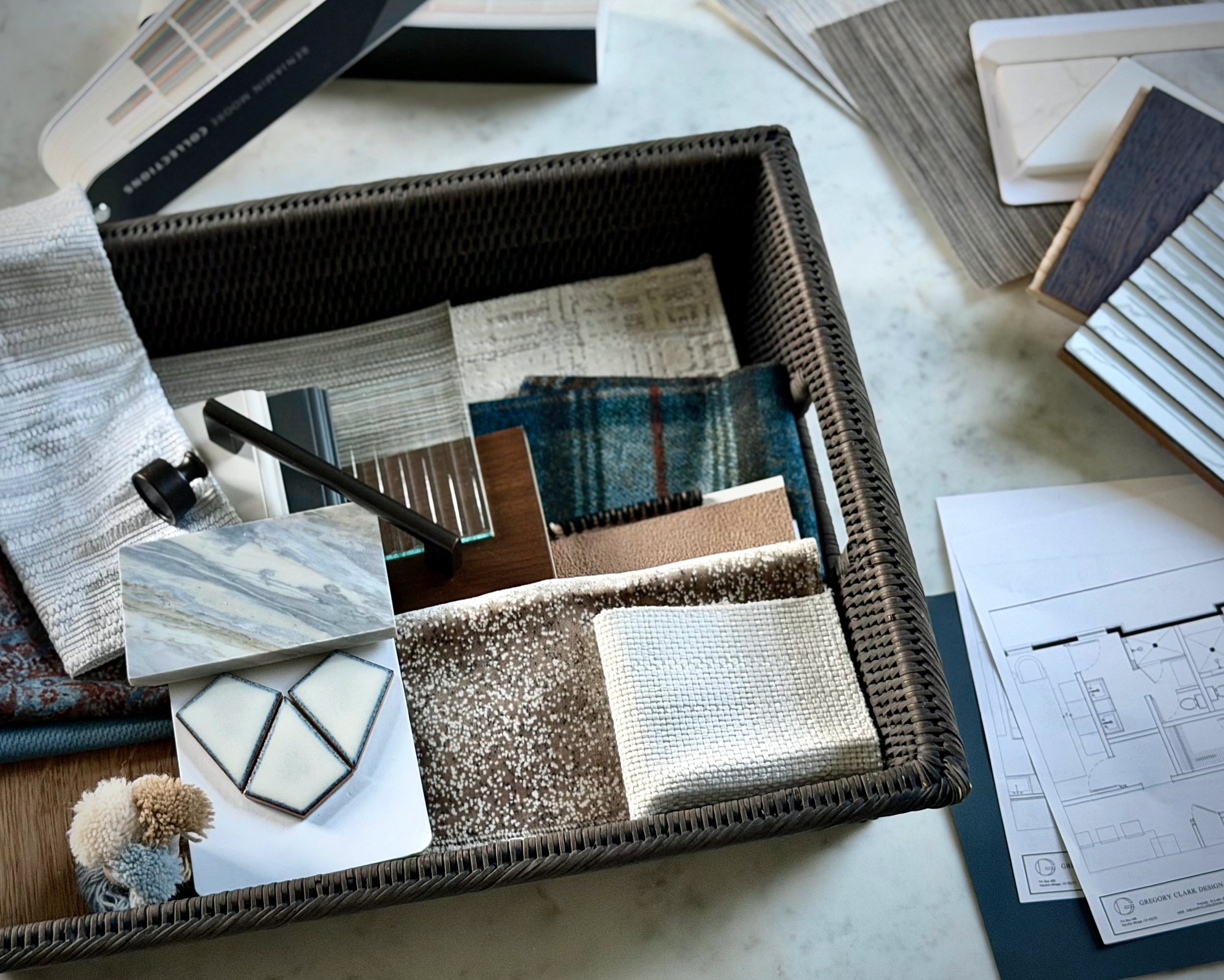 A display table with a custom high-end interior design palette of wallpaper samples; tile, countertop, flooring, cabinet finishes; hardware; paint colors; fabric and rug swatches; along with architectural blueprints and furniture floorpans.