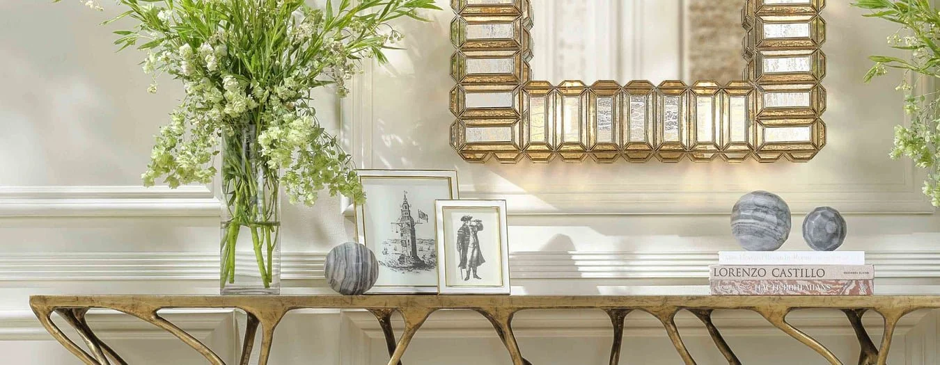 Elegant interior designer Made Goods room decor with a gold geometric mirror, a glass vase of white and green flowers, framed artwork, books, and decorative spheres on a wooden console table against a white wall.