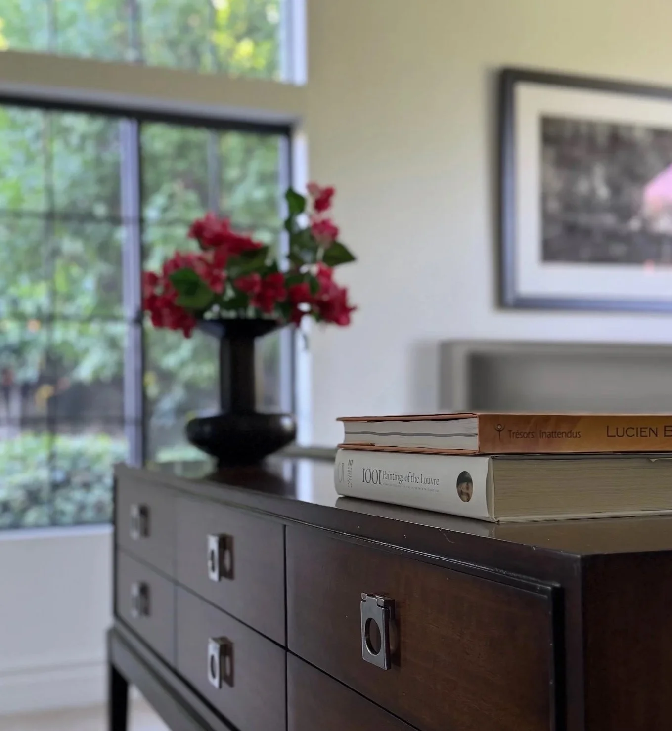A designer living room with mahogany wood console with drawers and silver handles, on top of which are books and a wood vase of pink flowers bougainvillea near a large window with greenery outside, framed picture on the wall, limestone fireplace.